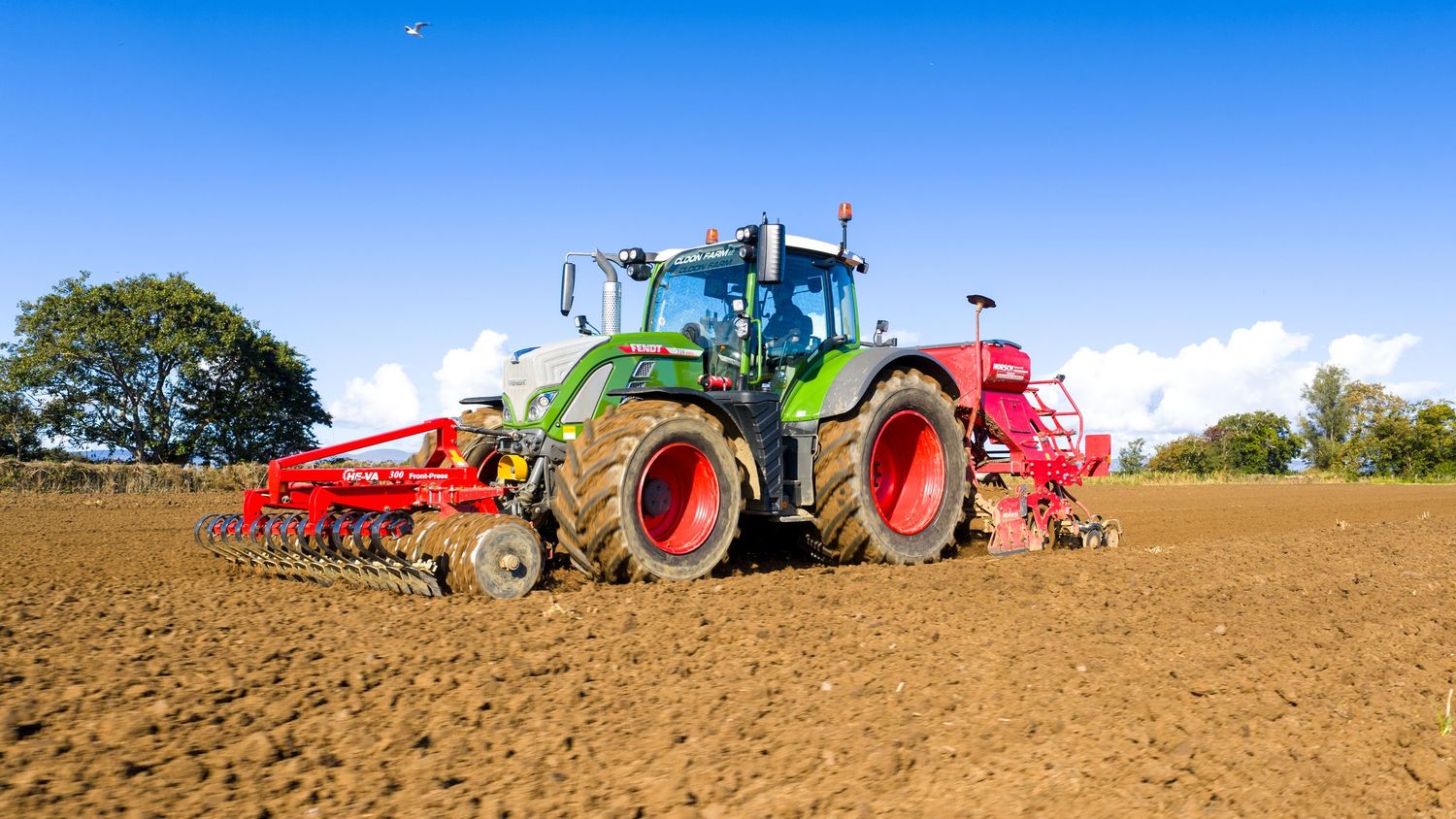 Green tractor with red agricultural equipment plowing a brown field under blue sky with white clouds.