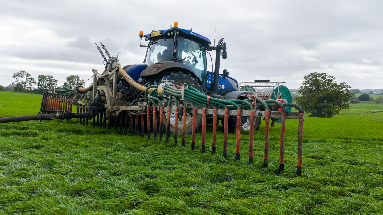Blue tractor with agricultural equipment spreading fertilizer on lush green farmland under cloudy skies.