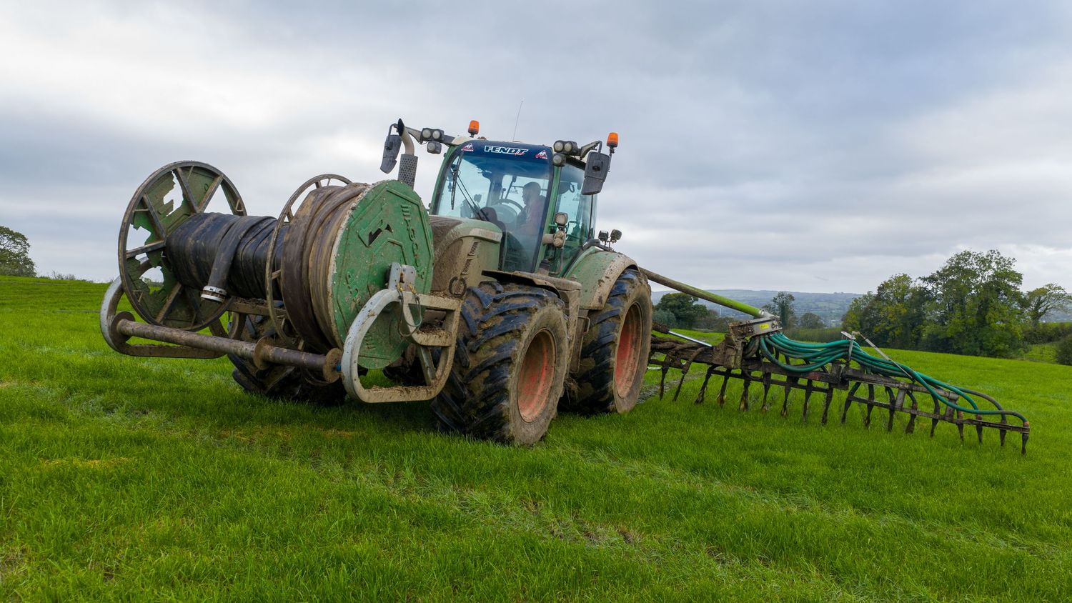 A panoramic view of agricultural machinery and tractors working in lush green farmland under cloudy skies.