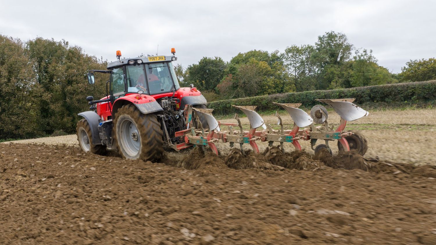 A red tractor pulling a plow creates neat furrows while cultivating agricultural soil in a rural field.