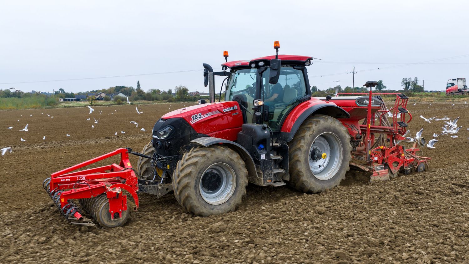 Red tractor pulling agricultural equipment across a plowed field with seagulls following behind.