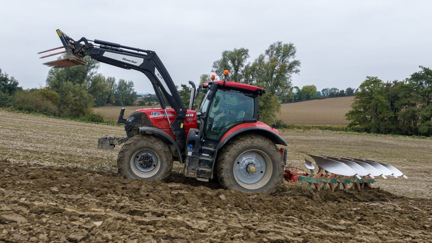 A tractor pulling a plow attachment creates straight furrows in a brown agricultural field during soil preparation.