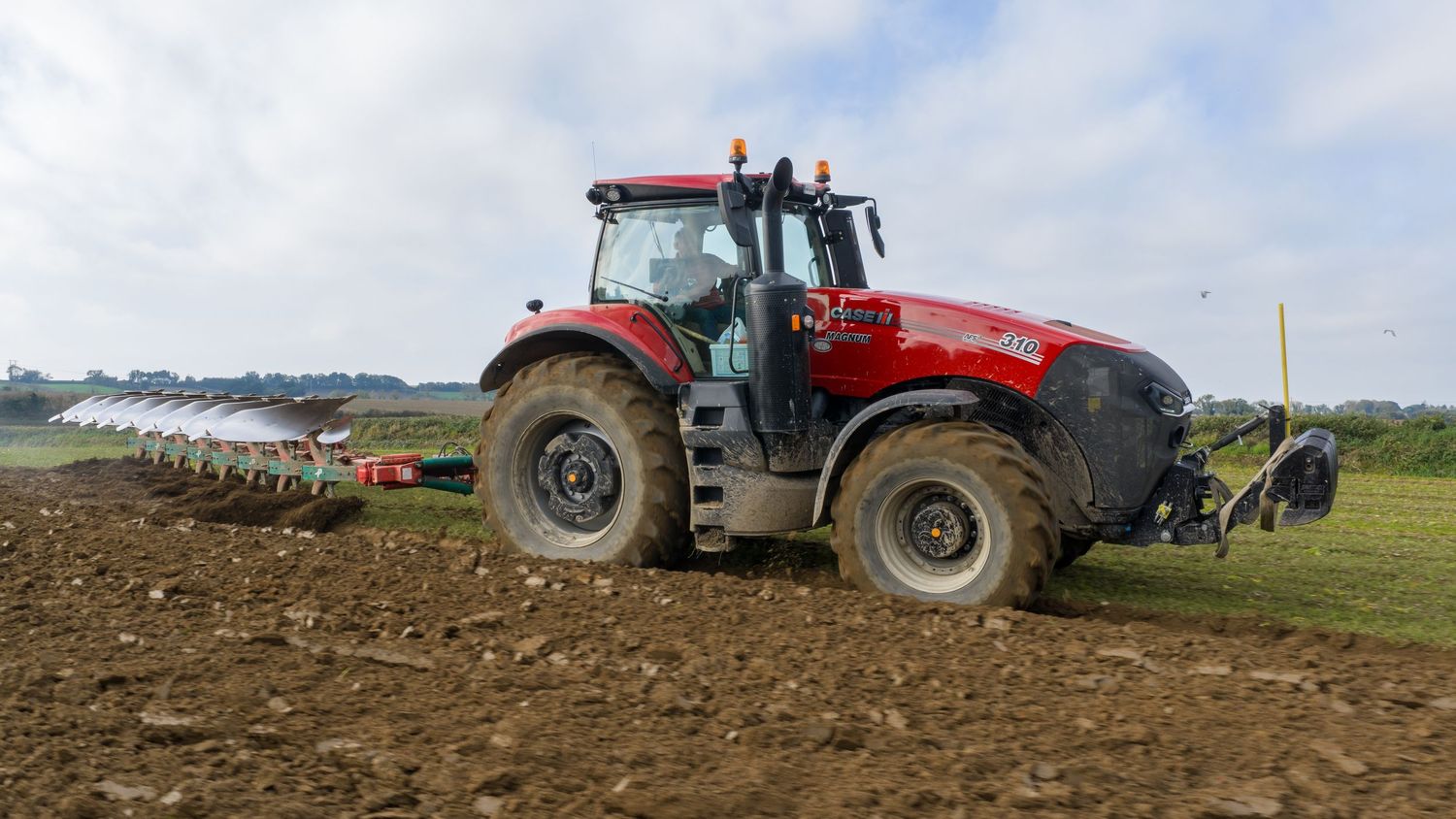 A red tractor plowing and cultivating agricultural fields against a cloudy sky in the countryside.