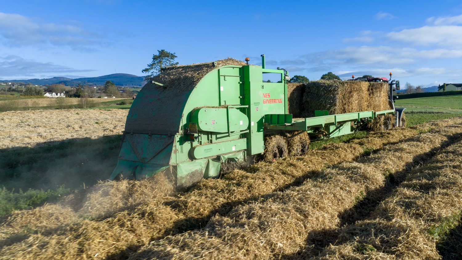Green agricultural harvesting machine working in a dry field with mountains visible in the background under blue sky.