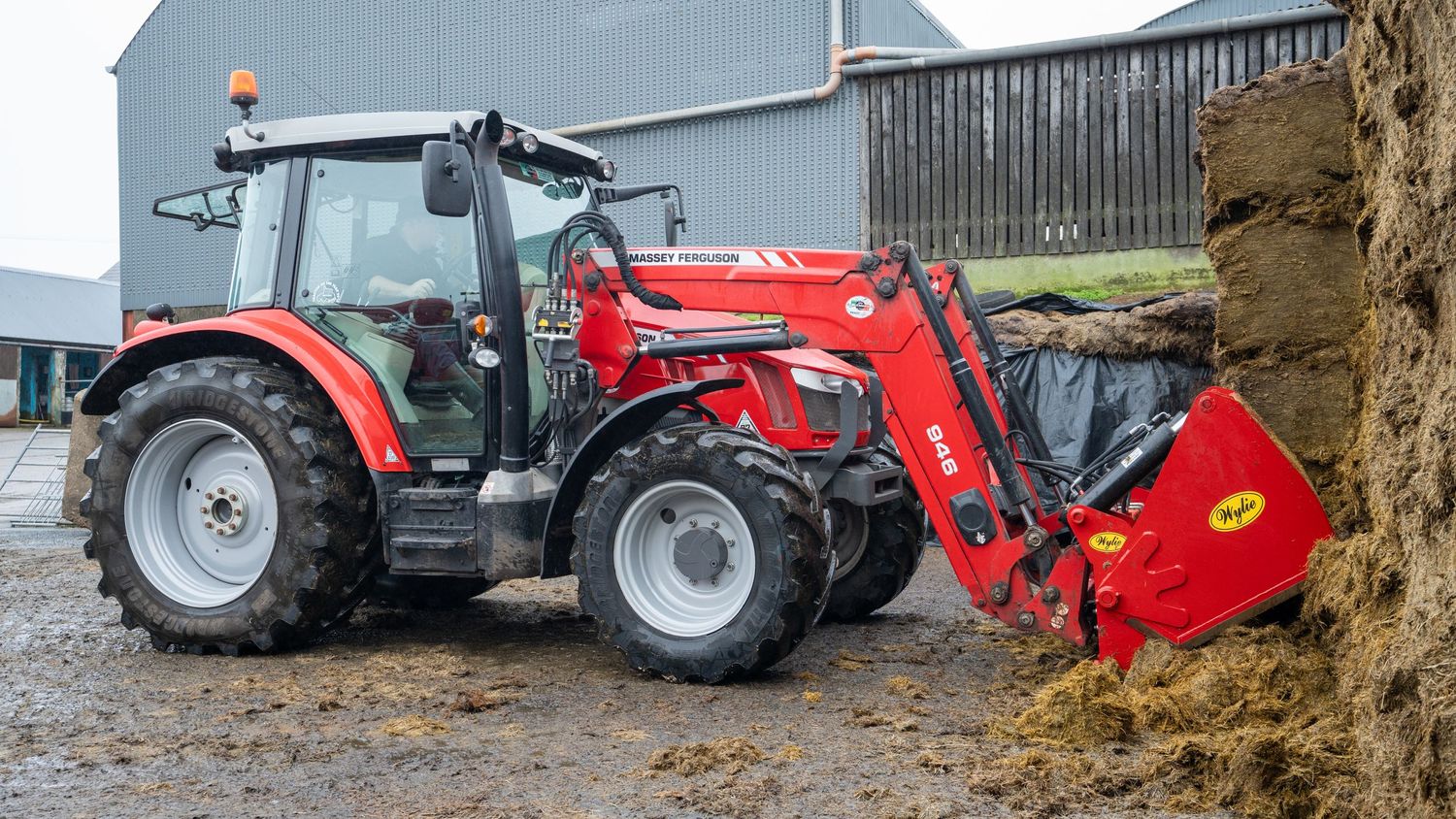 Red Massey Ferguson tractor with front loader attachment scooping material at a farm building site.