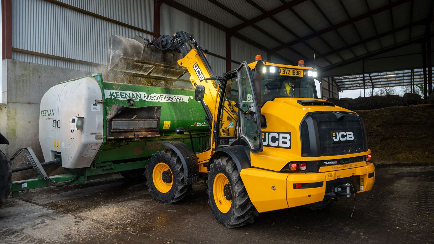 JCB compact wheel loader operating alongside farm equipment inside a storage shed.