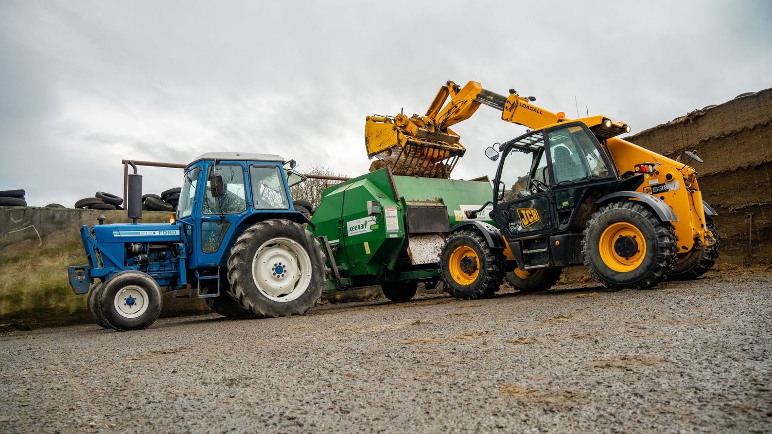 Blue vintage tractor and yellow telescopic forklift loading a green agricultural machinery on rural gravel road.
