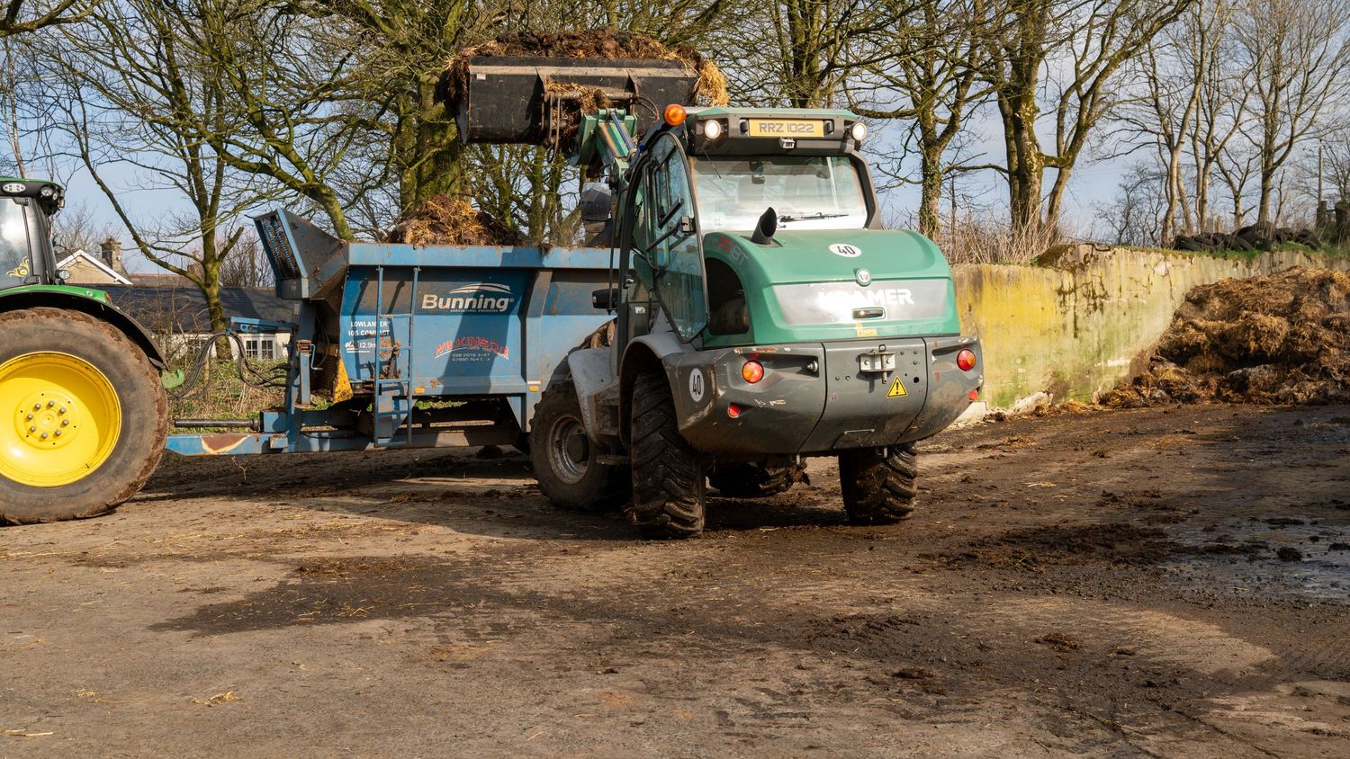 A green work truck with a dump bed parked on muddy ground next to farm equipment and trees.