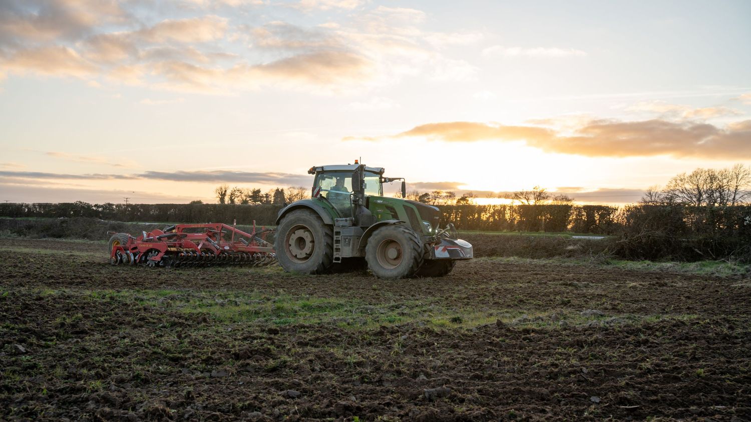 A tractor plows a field against a golden sunset sky with farm equipment silhouetted on the horizon.