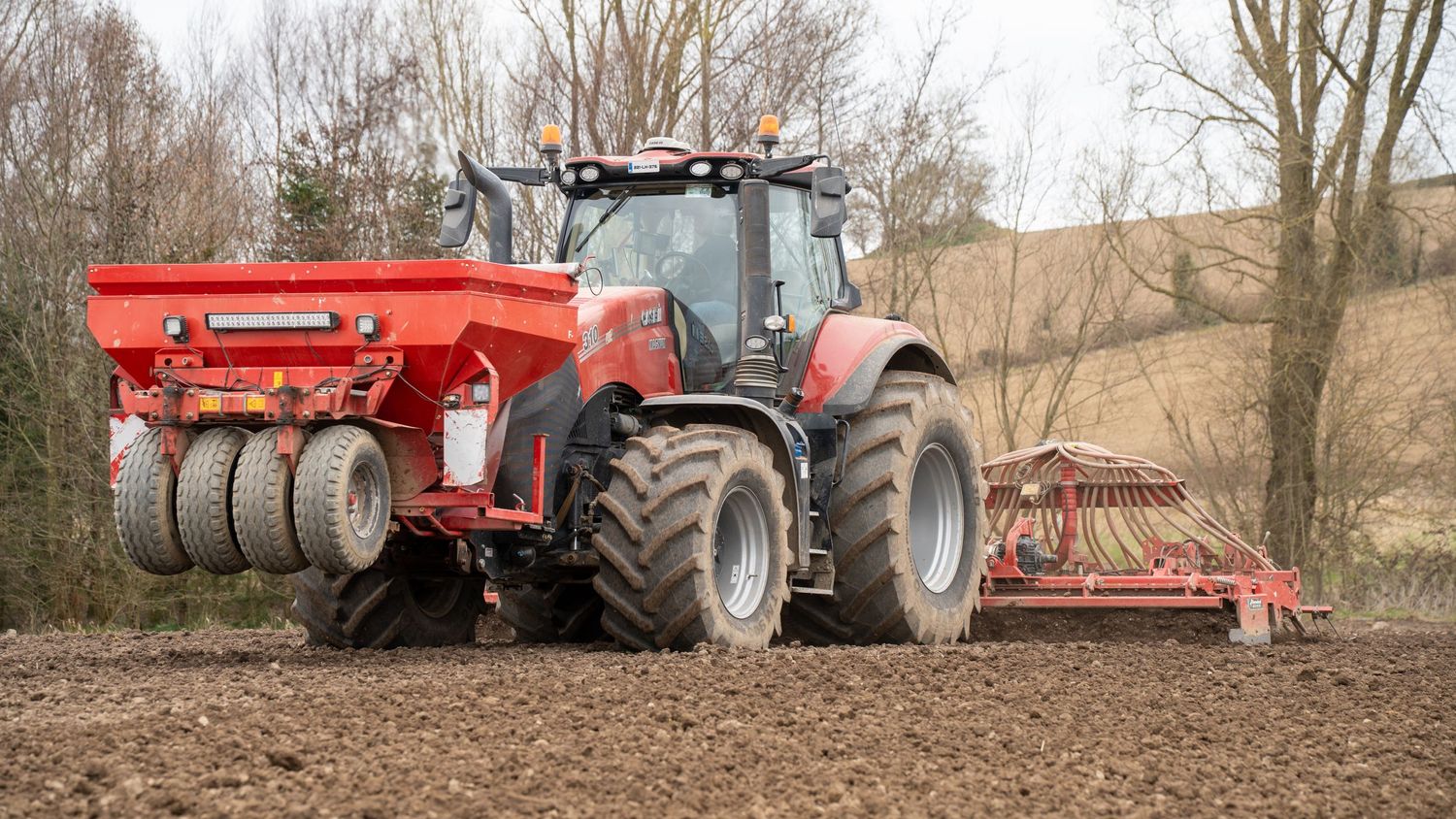 Large red tractor with fertilizer spreader attachment working in a plowed agricultural field on a cloudy day.