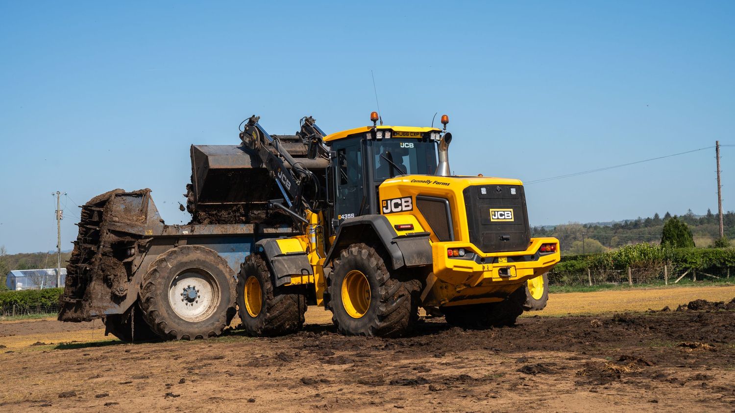 Yellow JCB wheel loader operating on dirt ground with agricultural equipment in background during daytime.