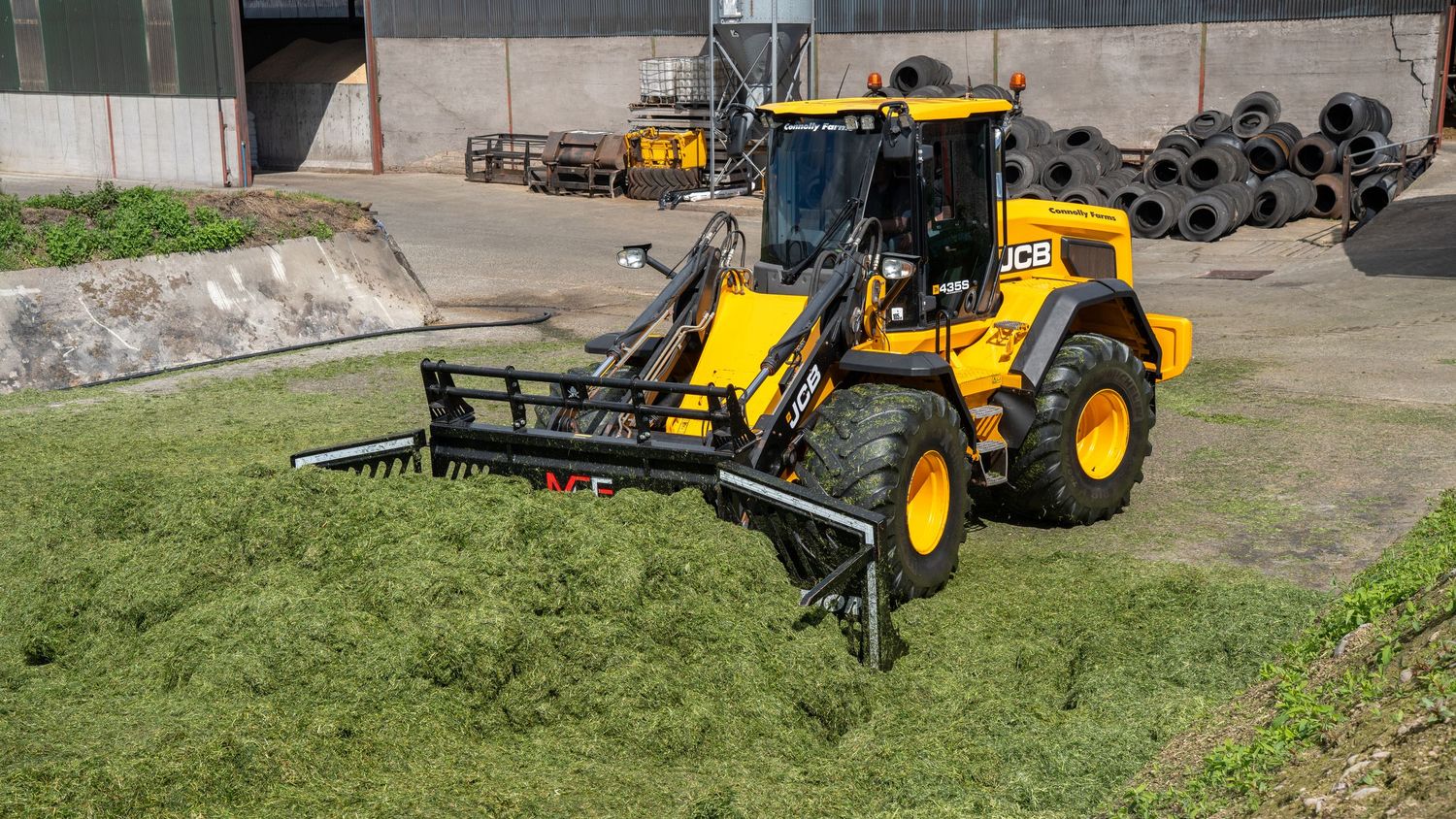 A yellow JCB compact wheel loader uses its front attachment to move piles of green silage or grass clippings.