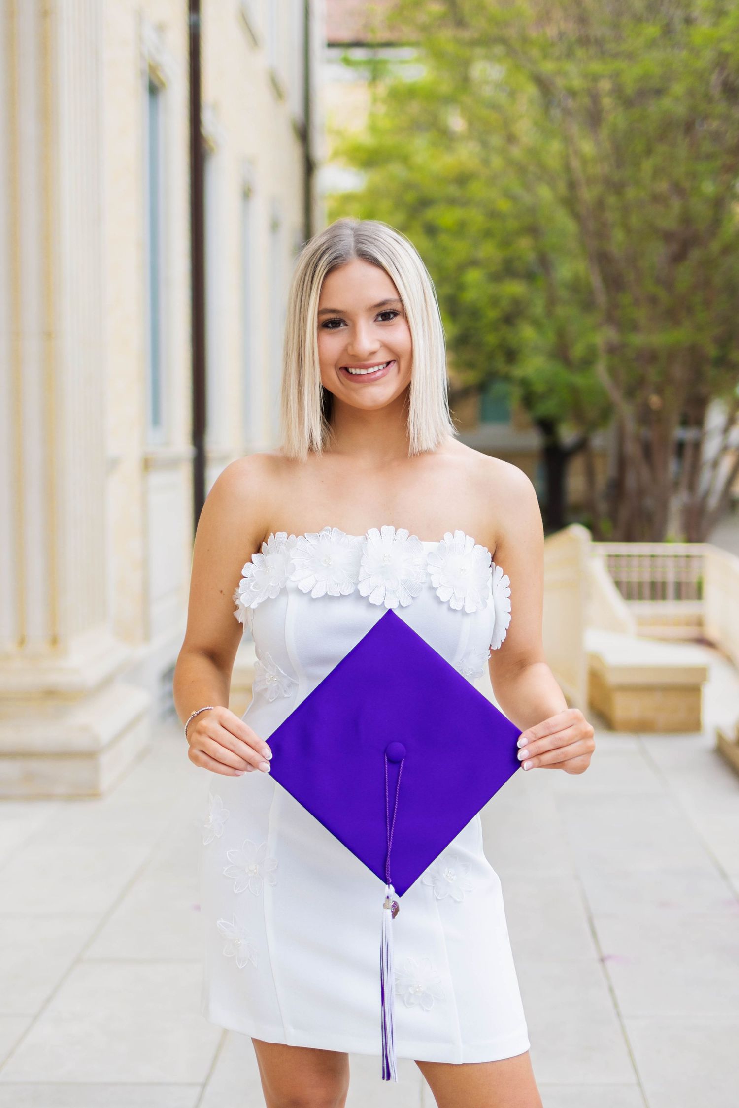 A graduate wearing a bright purple TCU graduation gown stands smiling on a university campus walkway.