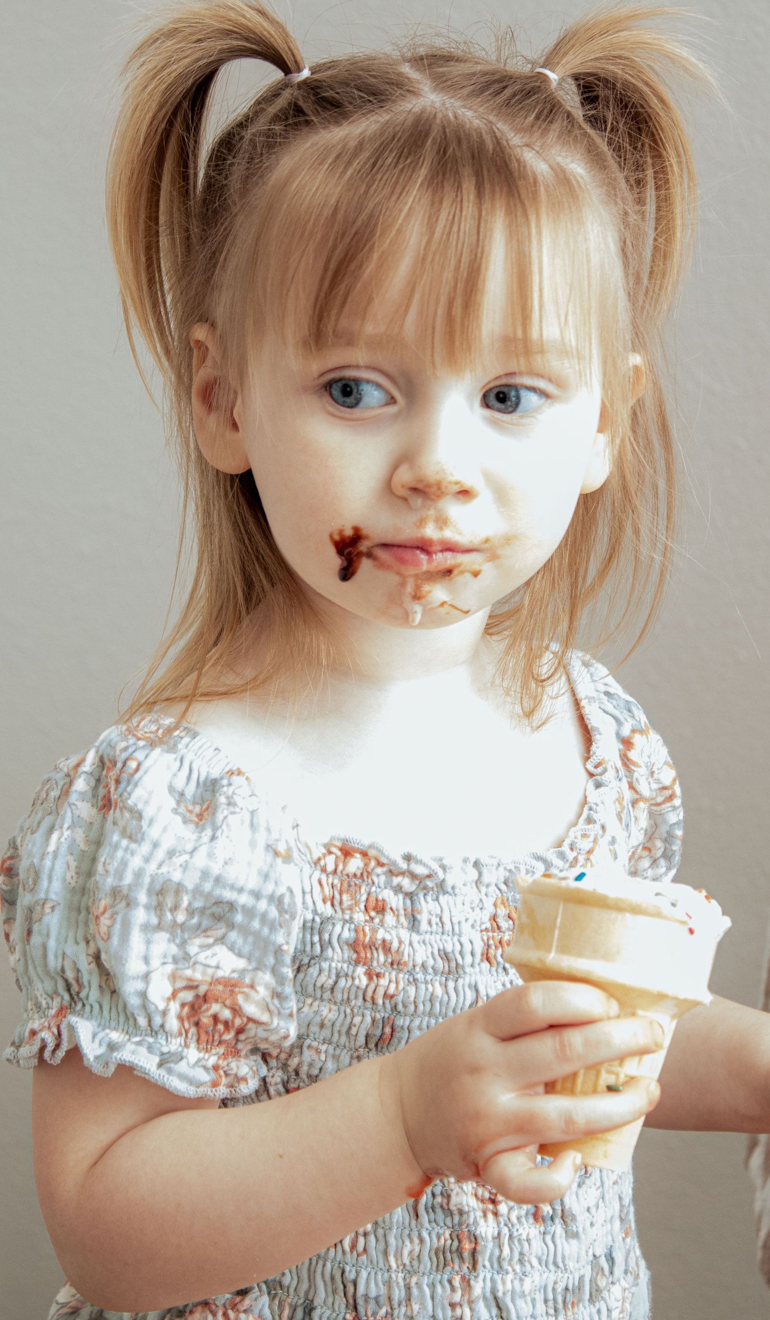 Small child in light floral dress holds an ice cream cone while wearing chocolate ice cream on their face.