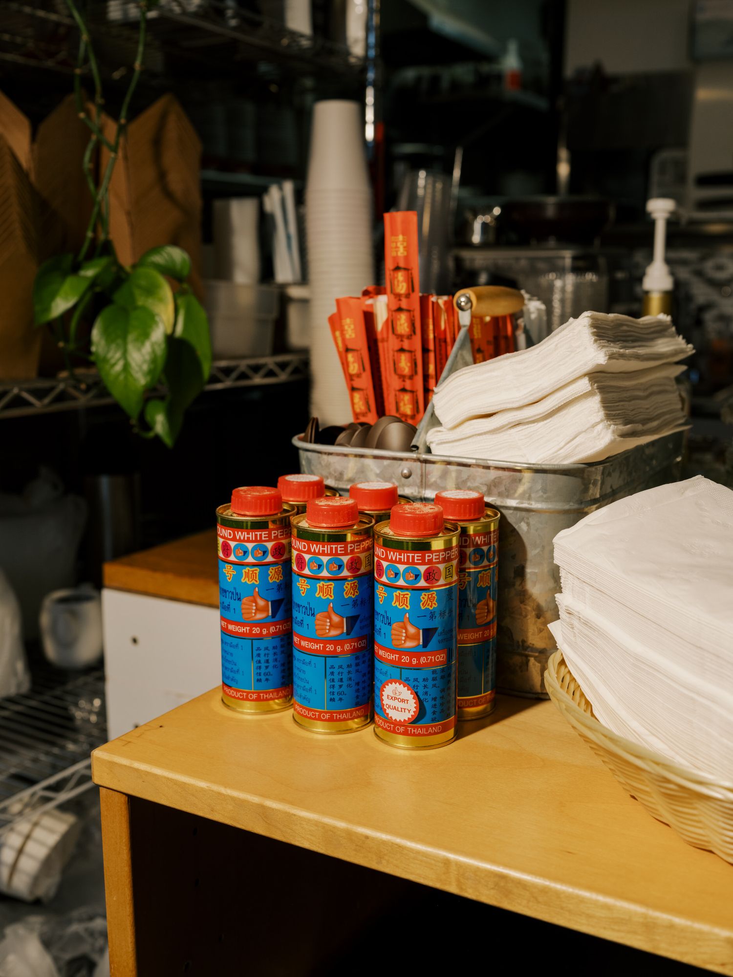 Multiple blue and orange Good Boy Cola cans stacked on a wooden shelf with towels and supplies in a restaurant setting.