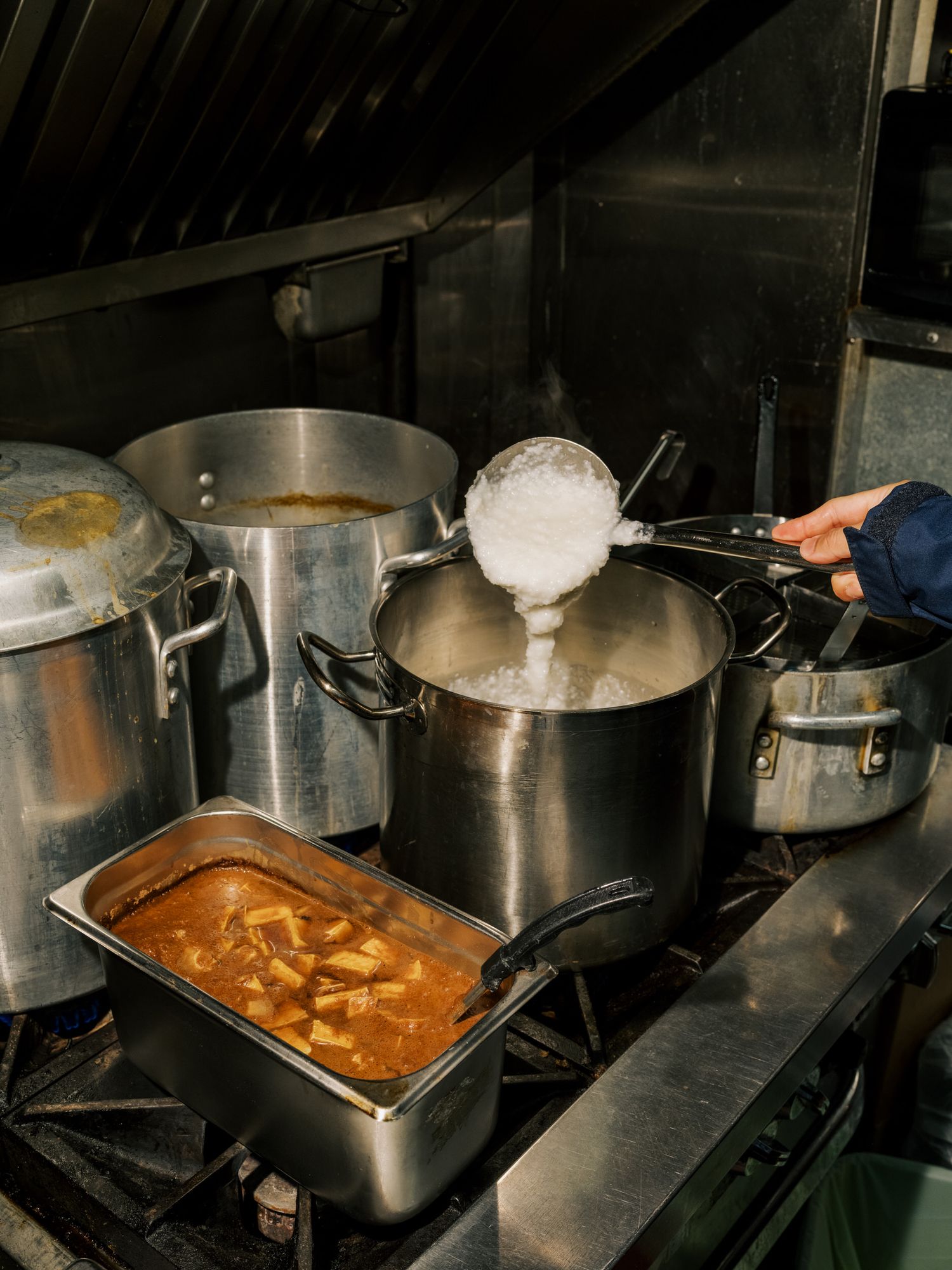Multiple stainless steel pots and pans cooking on a commercial stove with curry sauce and white mixture being prepared.