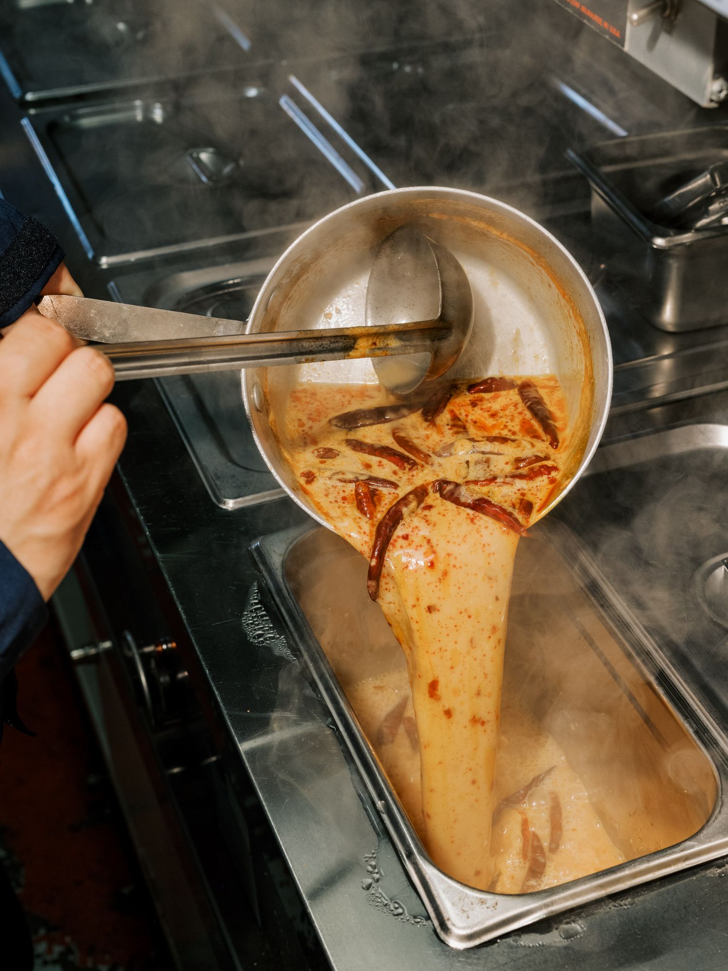 A hand pours a spicy kimchi sauce mixture from a steel bowl into a metal storage container on a professional kitchen stove.