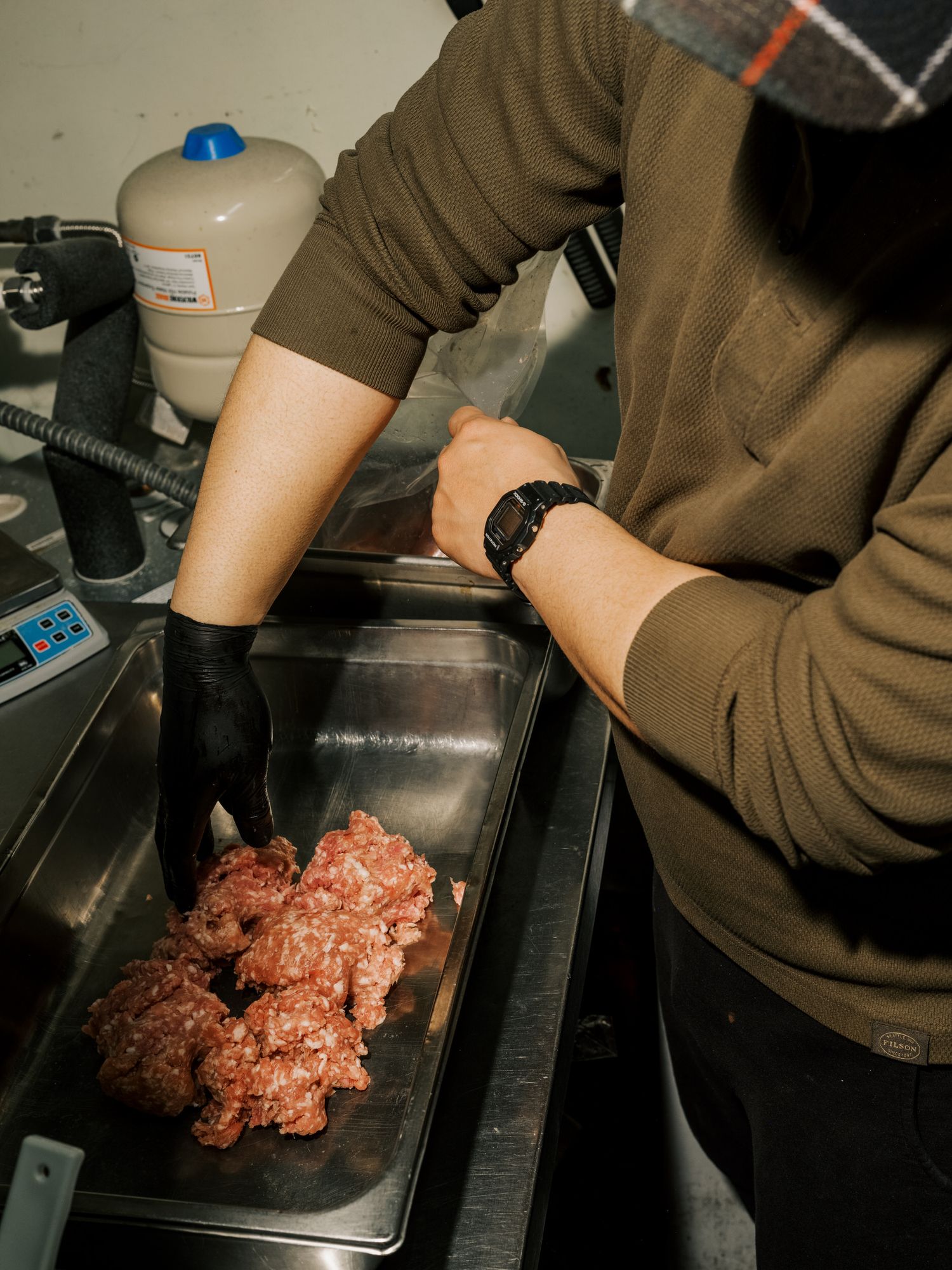Raw ground beef being prepared on a stainless steel surface by someone wearing a brown sweater and black watch.