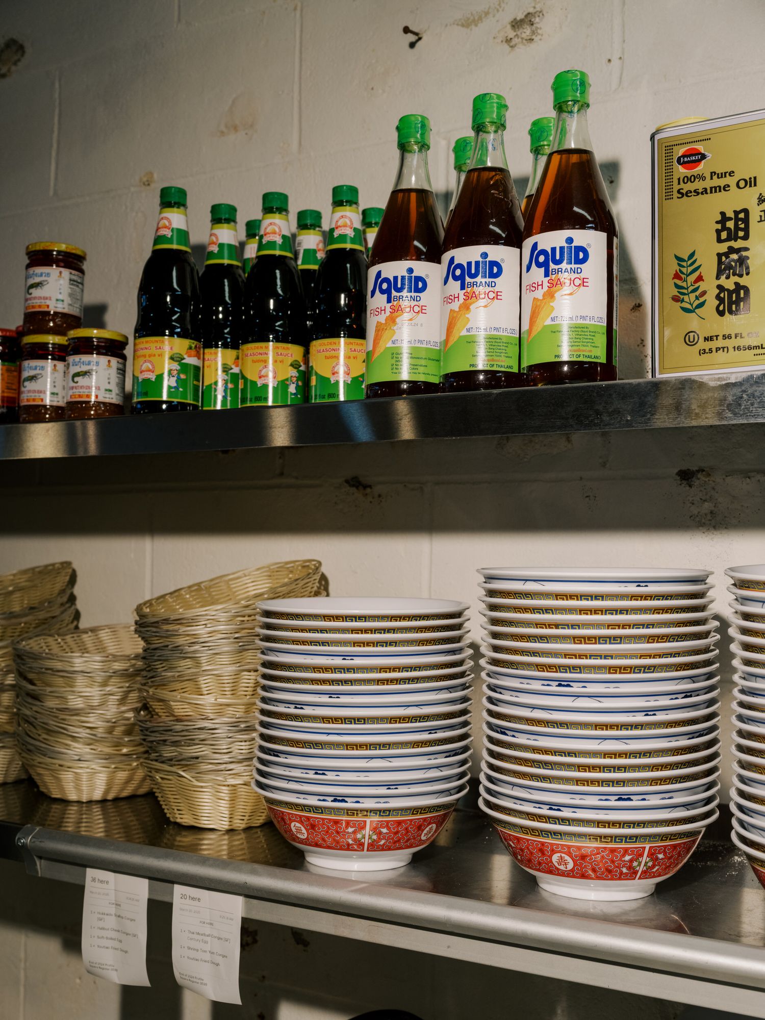 Stacked bowls and plates on metal shelving with green bottles of Sprite soda and Asian condiments in a restaurant storage area.
