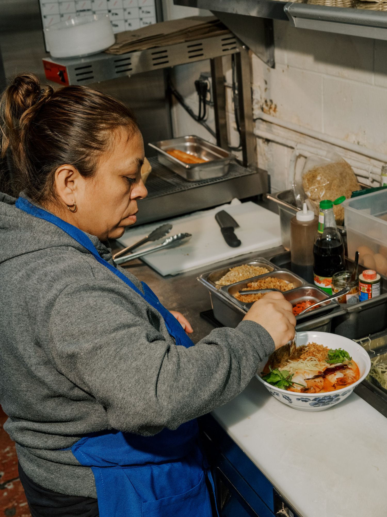 Kitchen worker in a gray sweater and blue apron preparing food at a restaurant counter with plates and ingredients.
