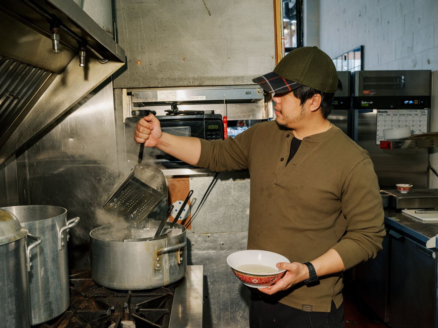 A cook in a brown sweater prepares food in a commercial kitchen while holding bowls near large stockpots.