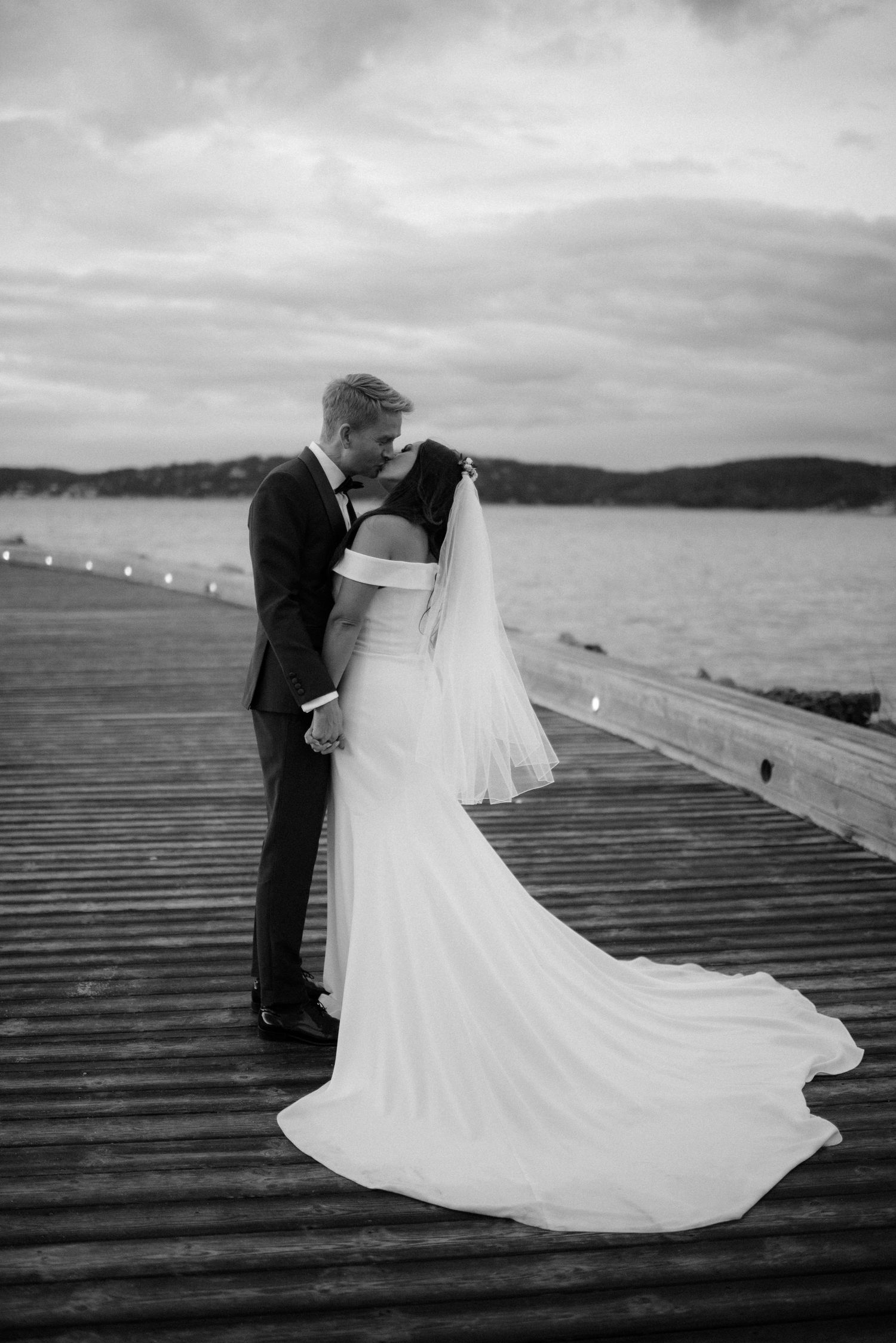 A black and white photo of newlyweds embracing on a waterfront dock with a long trailing wedding dress Villa Malla in Filtvet.
