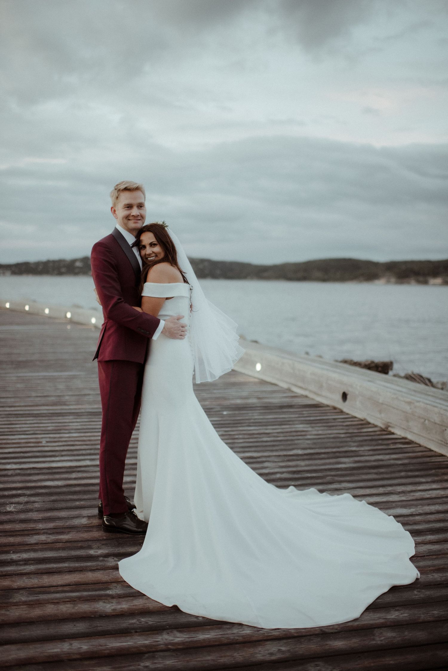 A bride in white gown and groom in burgundy suit share an intimate moment on a waterfront dock at dusk at Villa Malla in Filtvet, Norway.