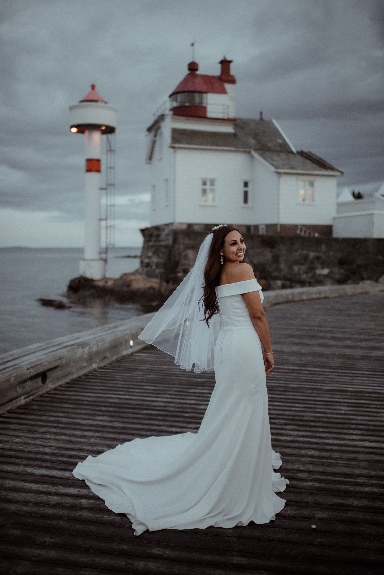 A bride in an elegant off-shoulder gown poses in front of a coastal lighthouse on a wooden pier at Villa Malla in Filtvet, Norway.