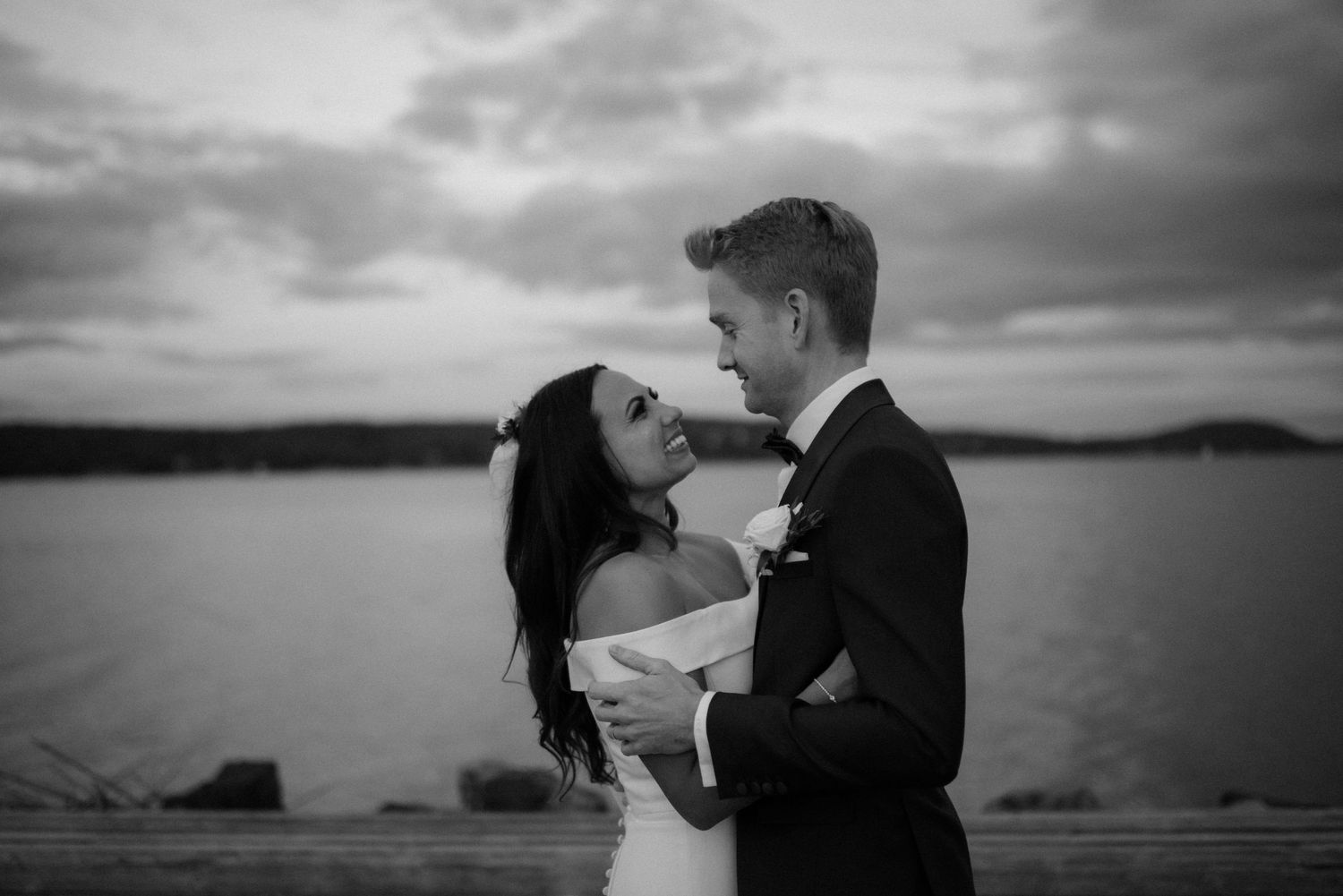 A romantic black and white wedding photo of a couple embracing at sunset by a calm waterfront at Villa Malla in Filtvet, Norway.