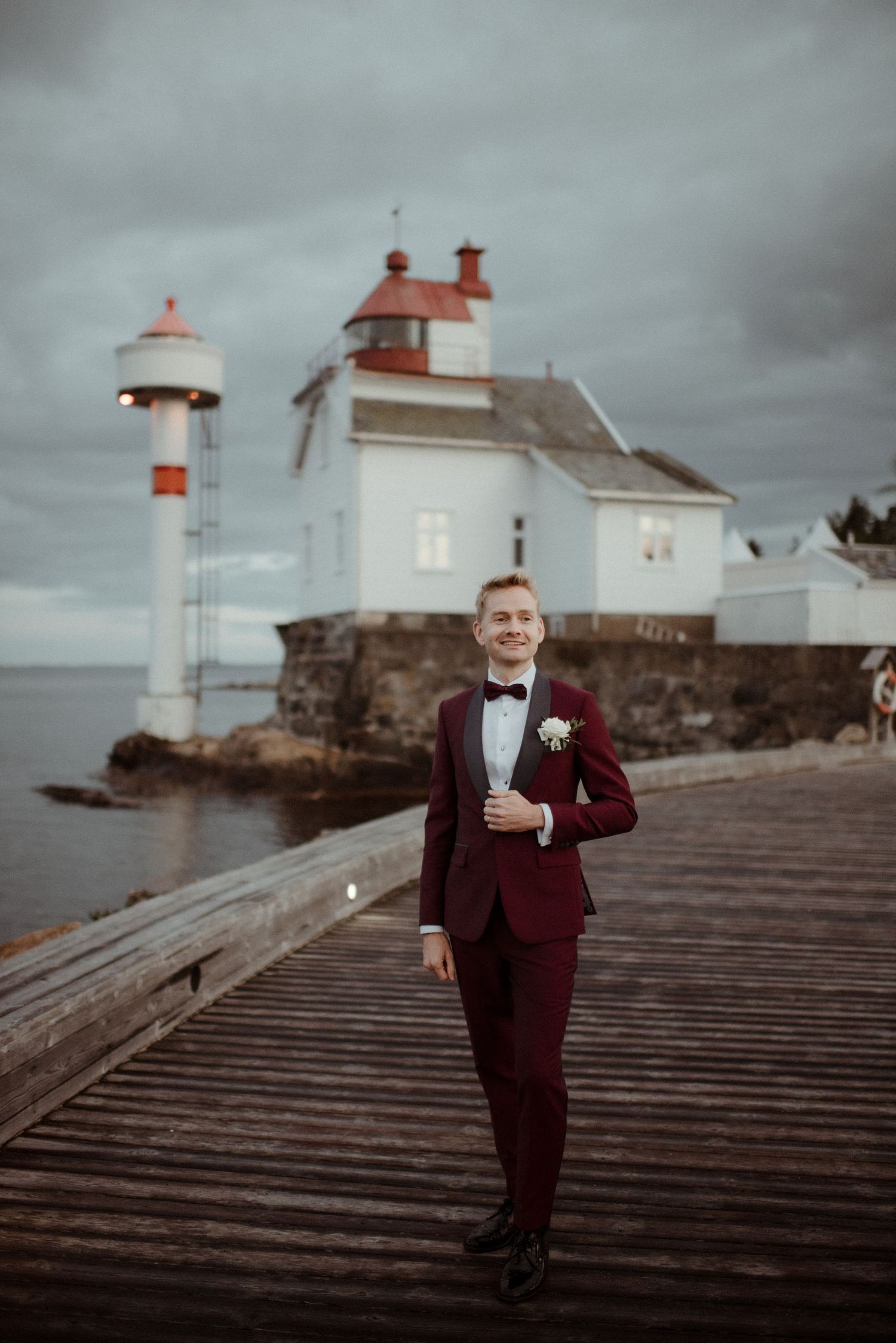 A burgundy tuxedo-clad figure poses on a wooden pier with a white lighthouse and stormy sky in the background at Villa Malla in Filtvet.