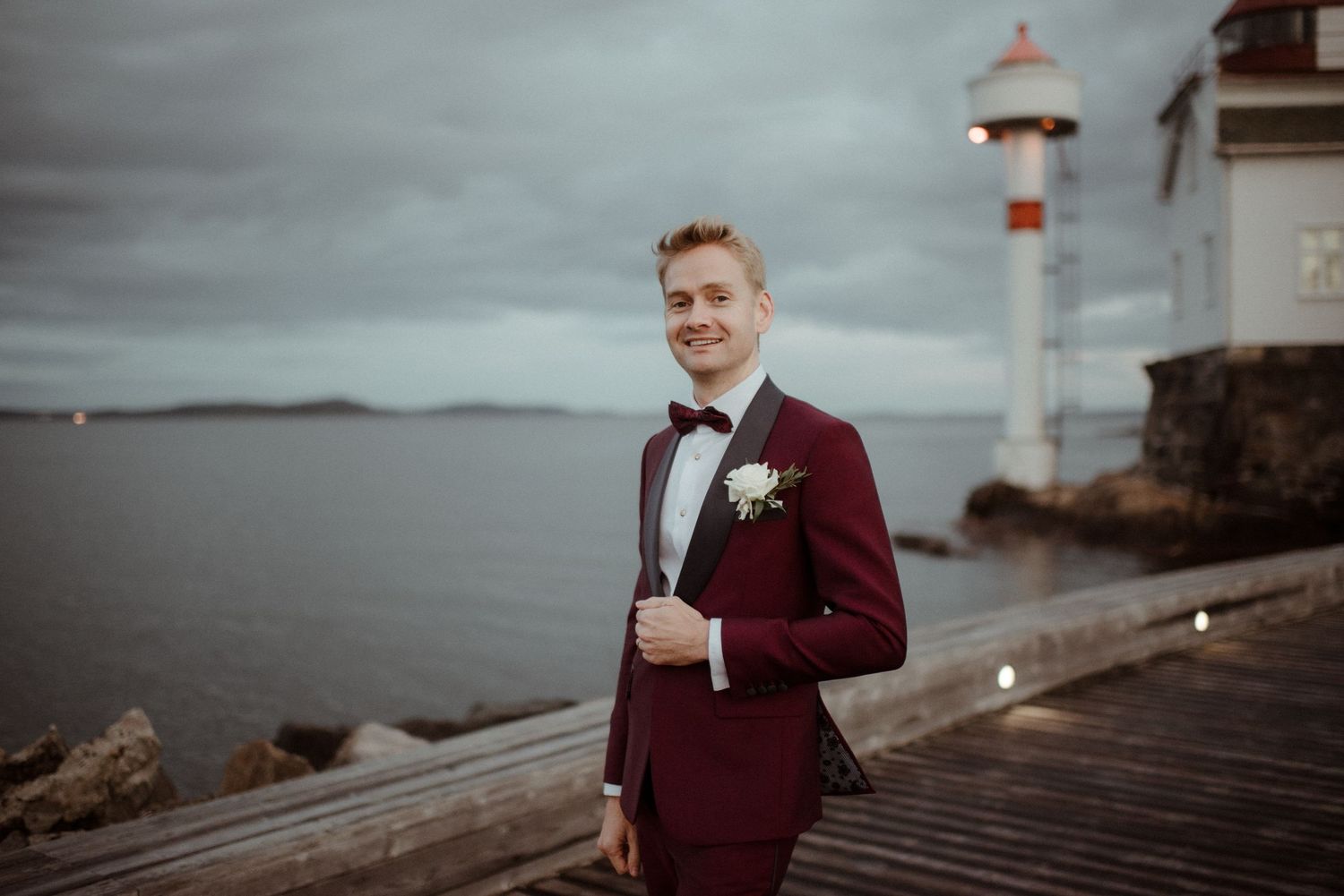 A person in a burgundy tuxedo stands on a waterfront boardwalk with a lighthouse in the background on an overcast day at Villa Malla.