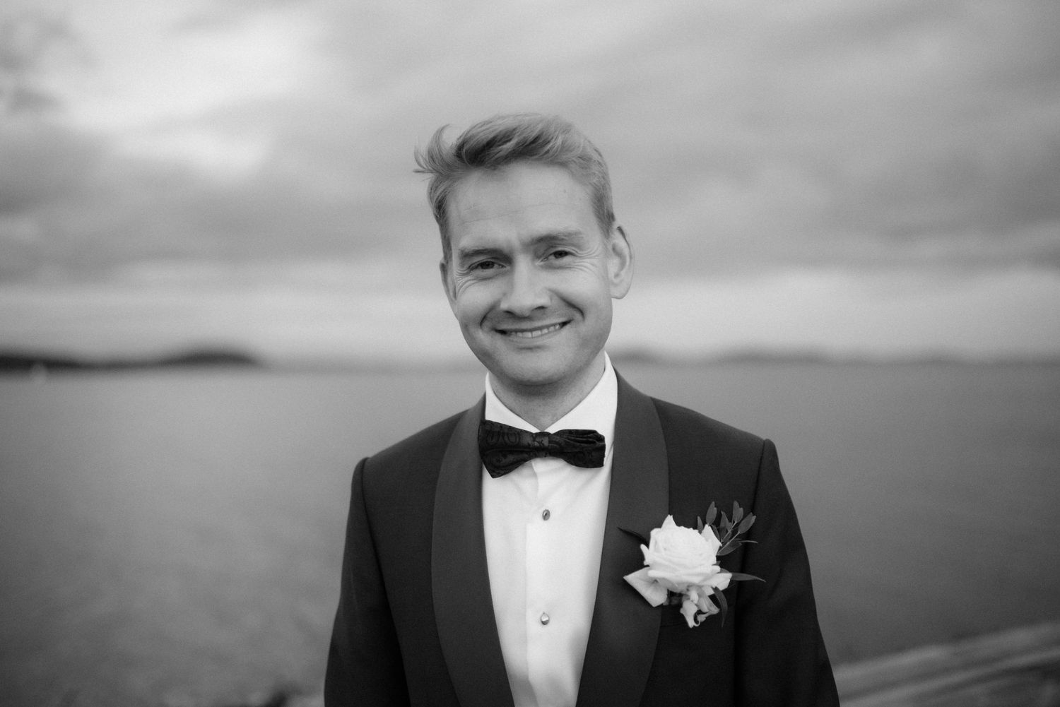 A person wearing a black tuxedo and bowtie smiles warmly at the camera in a black and white photograph by the fjord at Villa Malla in Filtv