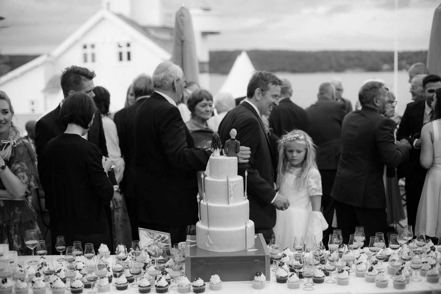 A black and white photograph of a wedding reception with guests gathered around a tiered white cake at Villa Malla in Filtvet, Norway.