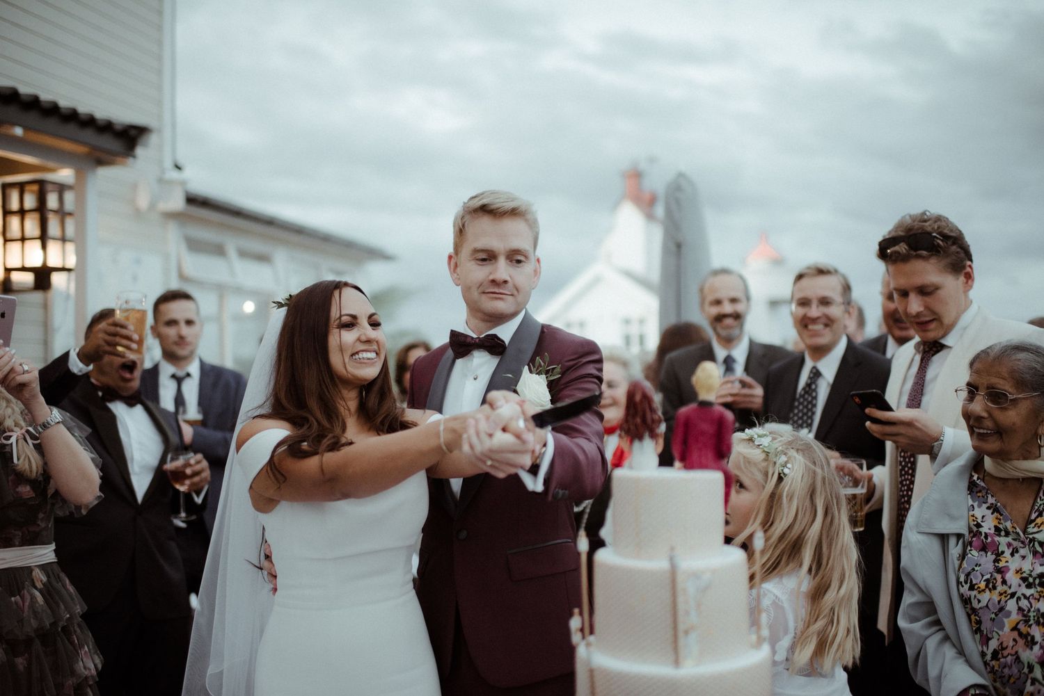 Wedding couple cutting their multi-tiered white cake surrounded by celebrating guests at Villa Malla in Filtvet, Norway.