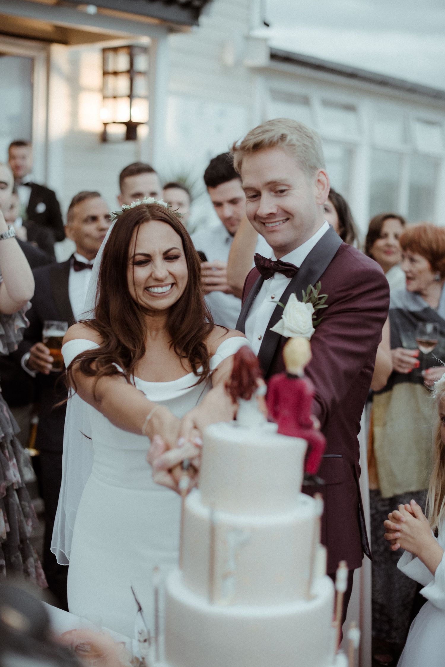 A wedding couple cuts their elegant white tiered cake surrounded by celebrating guests at their reception at Villa Malla in Filtvet, Norway.