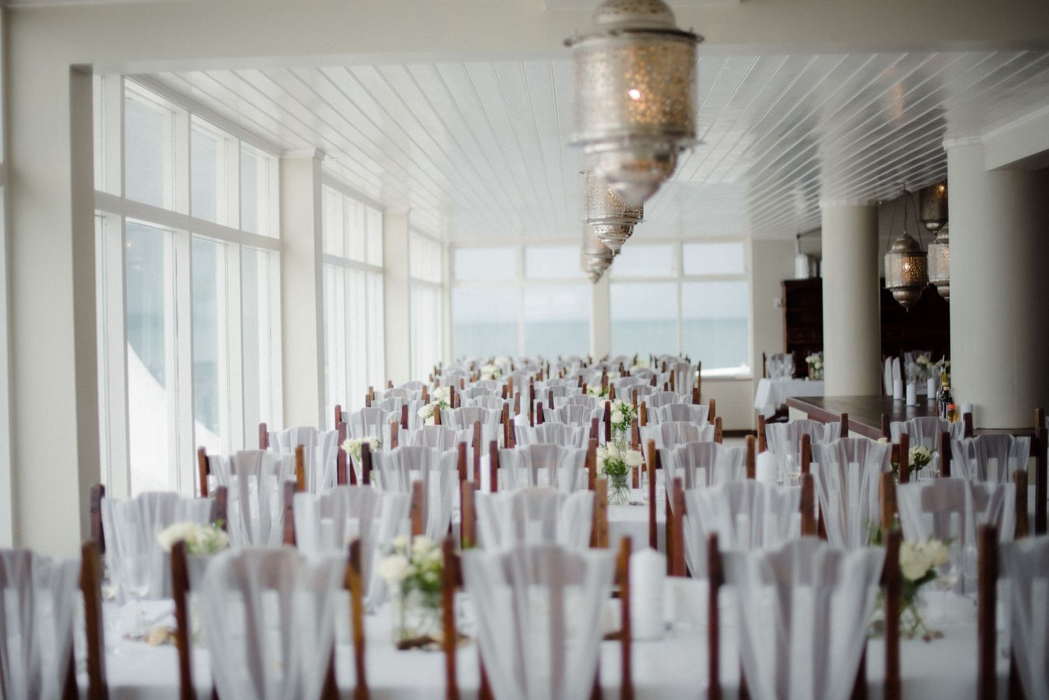 An elegant wedding reception setup with white chairs adorned with burgundy ribbons and greenery in a sunlit ballroom at Villa Malla.