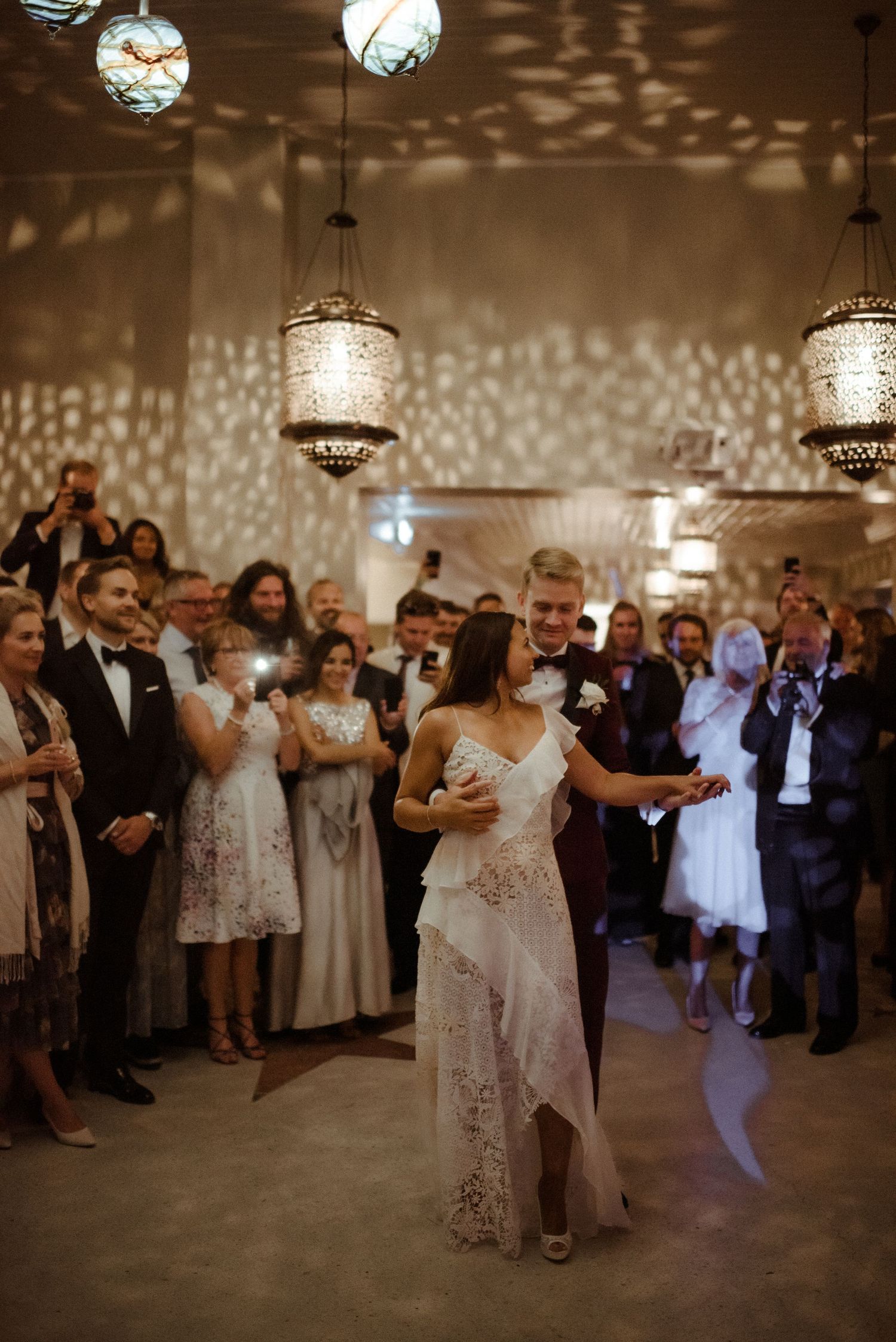 A couple shares their first dance at a wedding reception under ornate chandeliers casting intricate shadows on the walls.