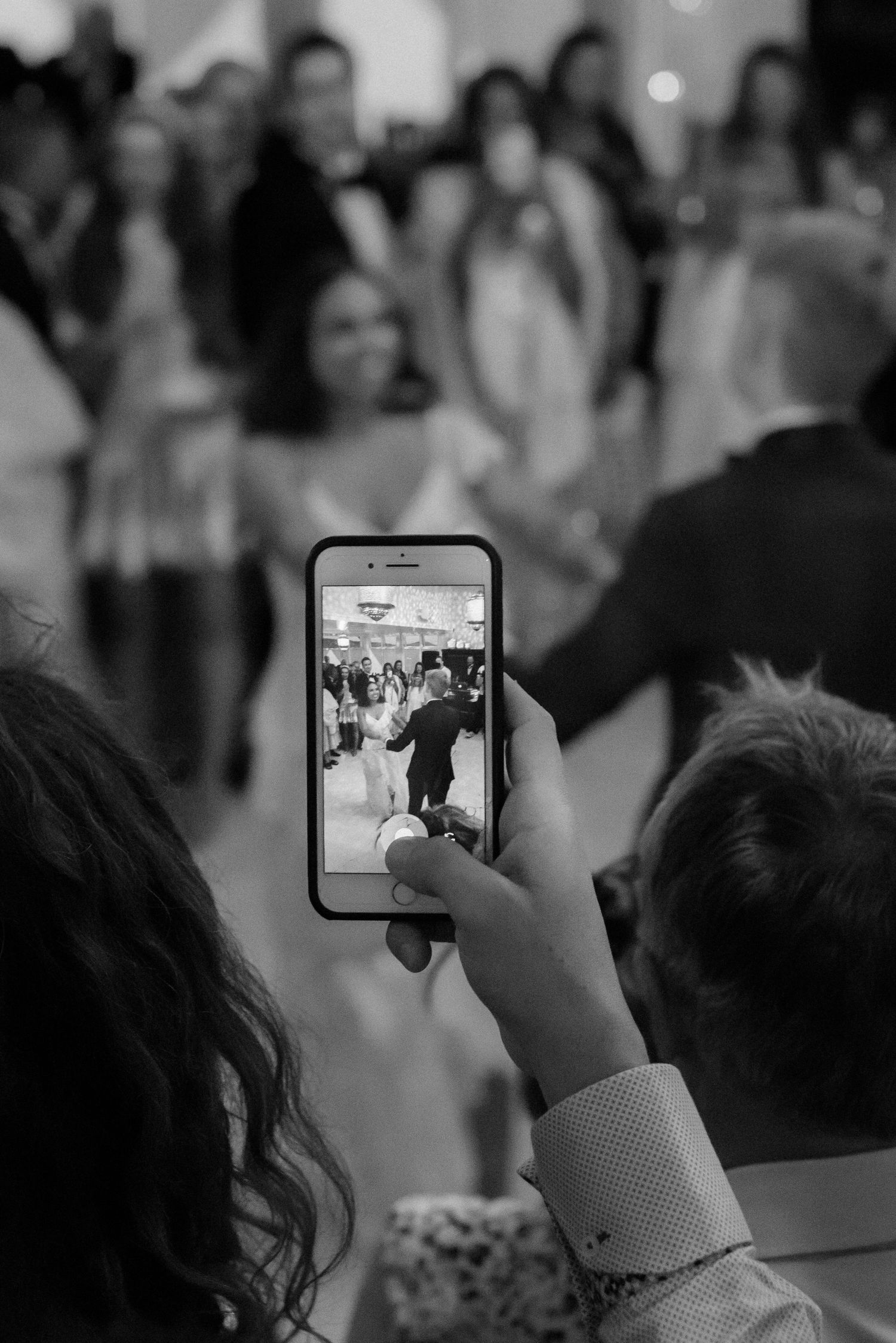 Someone holds up a smartphone to capture a dance moment at a wedding reception in black and white.