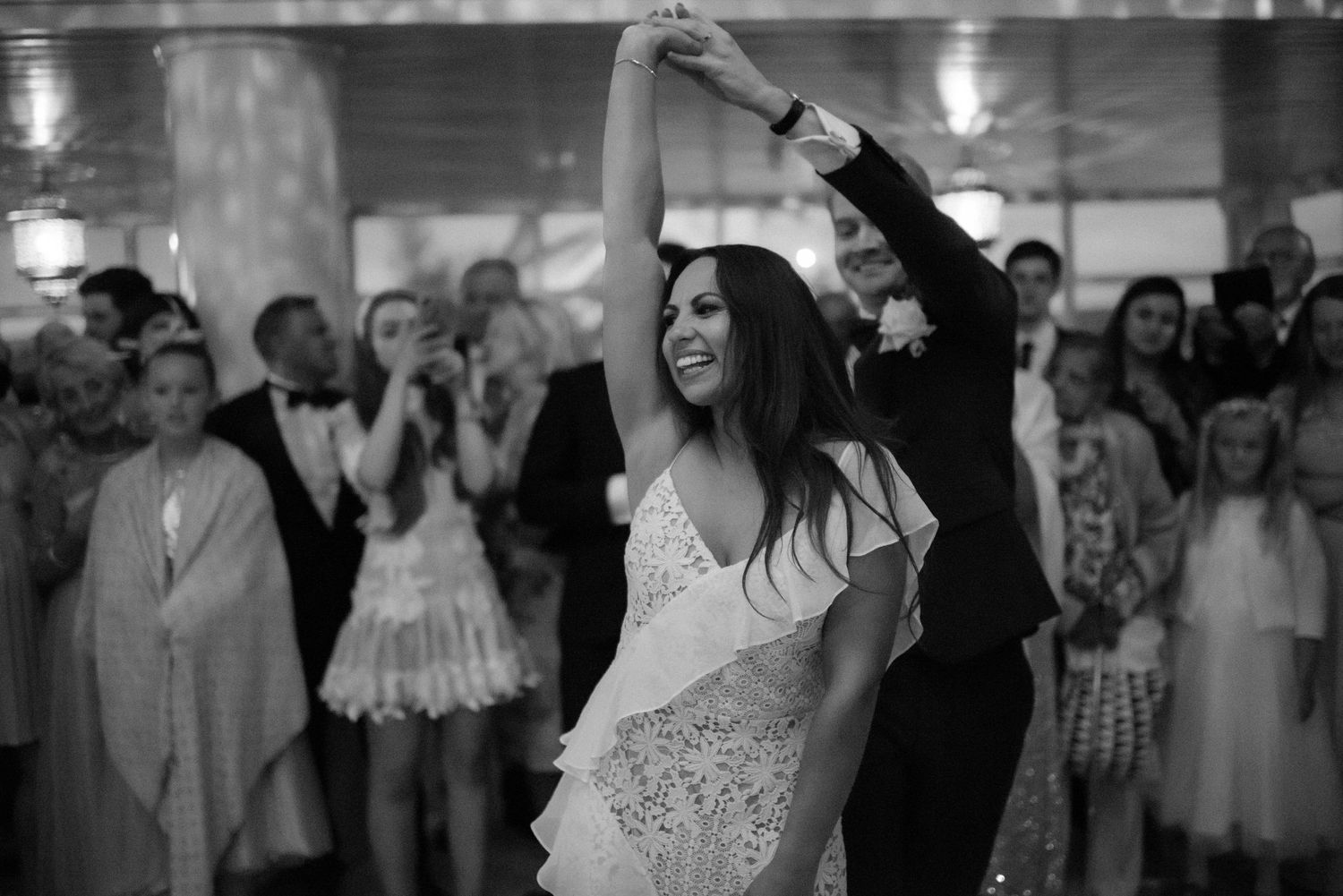 A couple dances enthusiastically in black and white at a wedding reception with guests watching in the background.