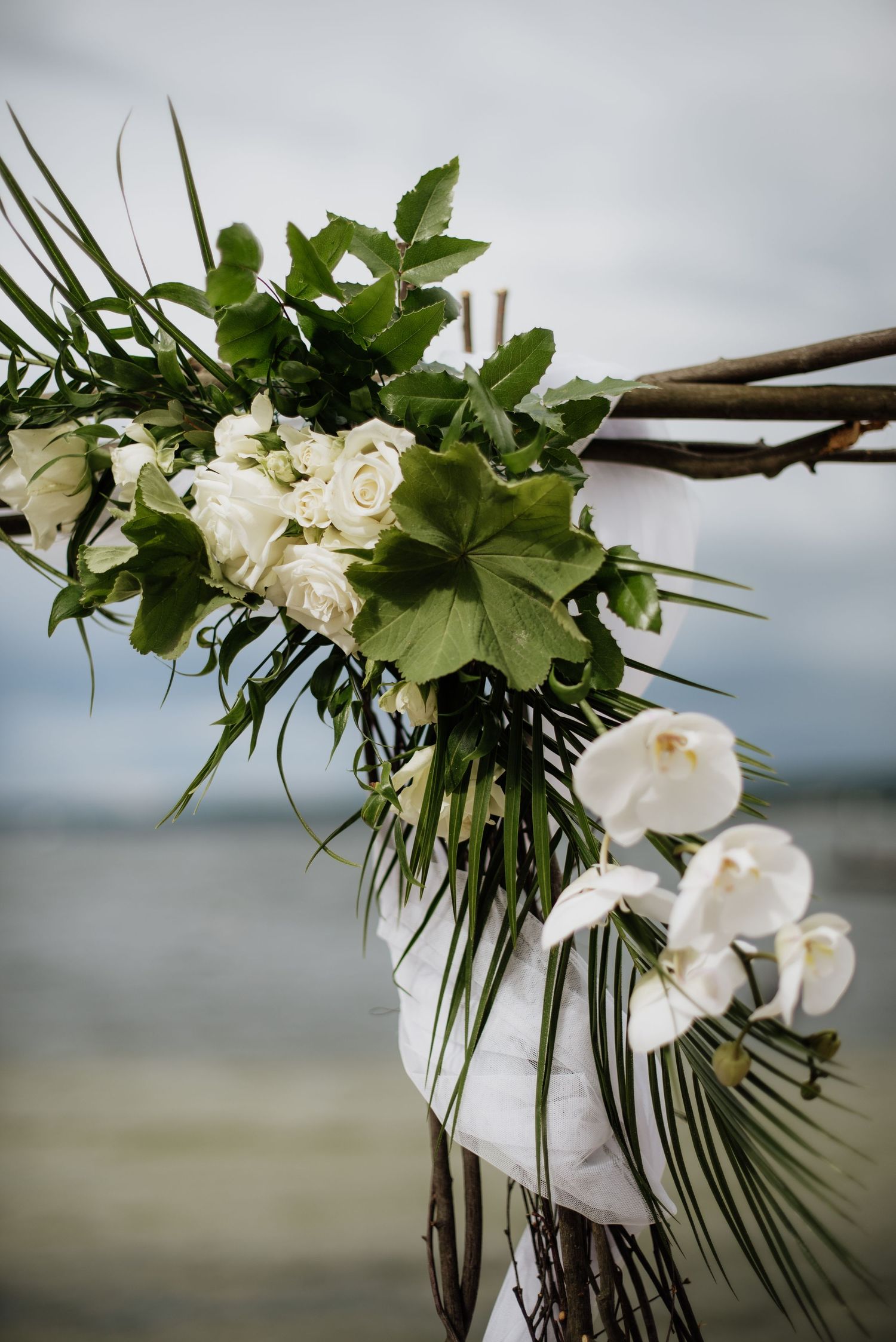 Elegant white roses and tropical greenery decorate a wooden wedding arch frame against a cloudy beach backdrop.