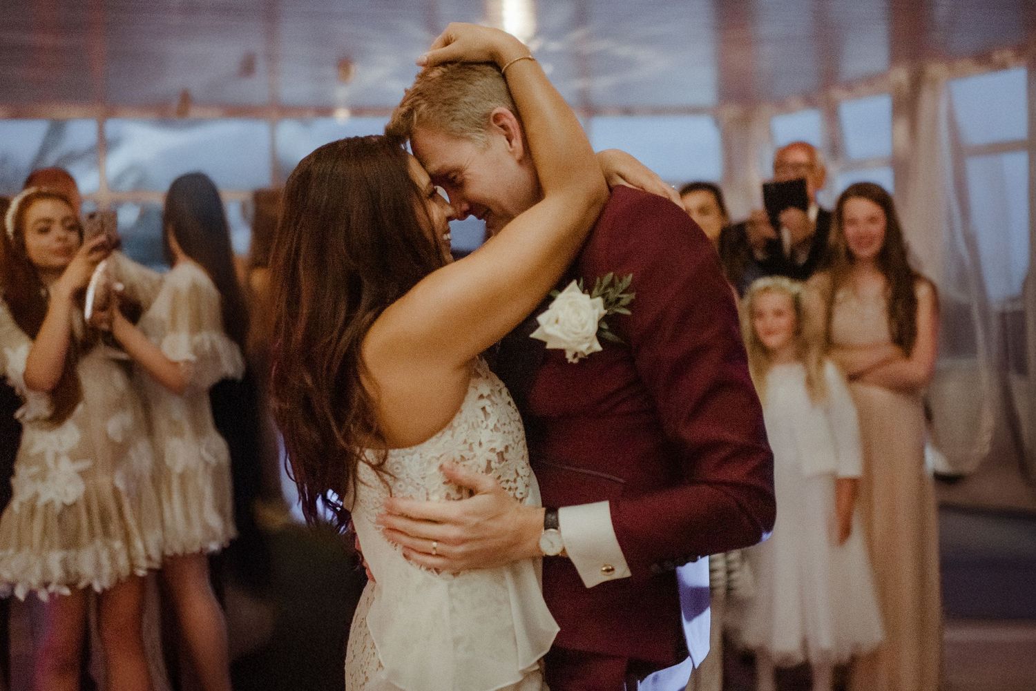 A bride and groom share a romantic first dance while wedding guests watch in the background of a dreamy reception venue.