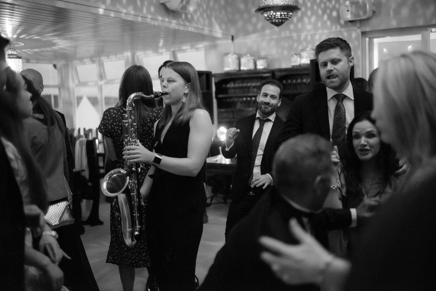 Black and white photo of people socializing at an elegant indoor party with crystal chandeliers overhead.