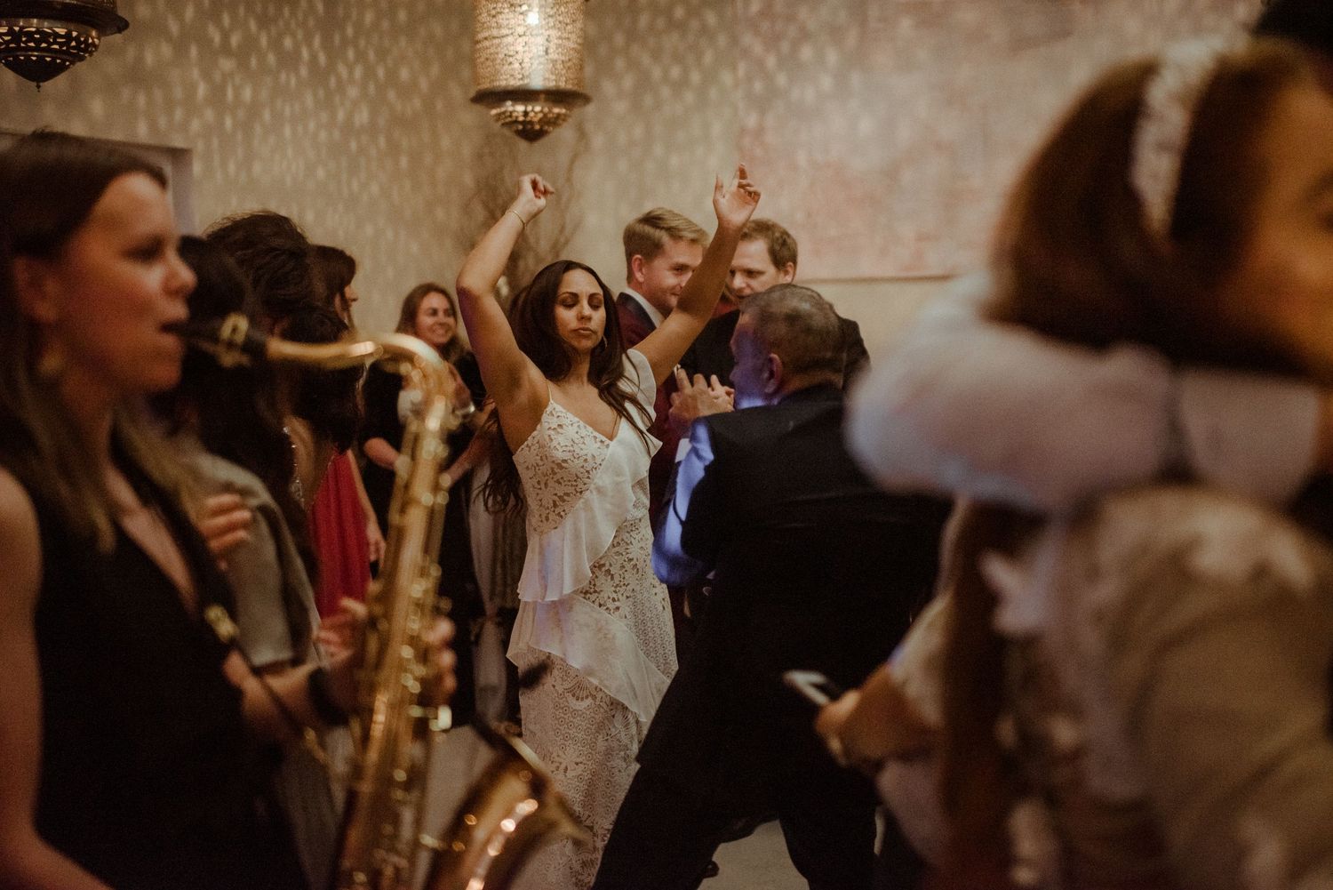 Wedding guests dance and celebrate on a dimly lit dance floor with a saxophone player visible in the foreground.