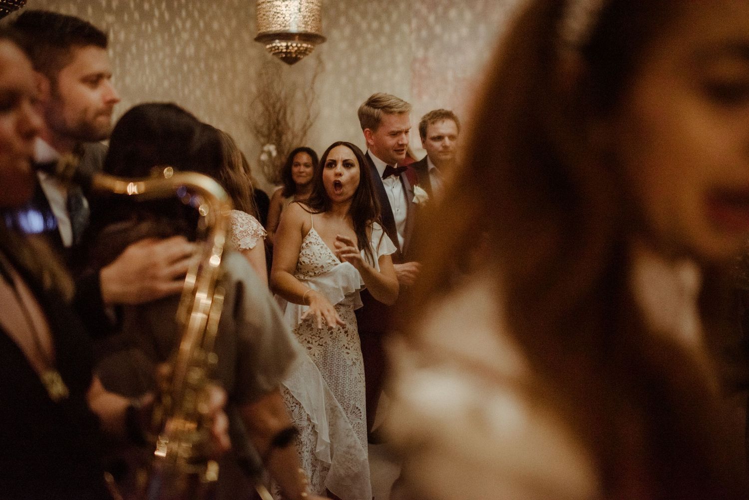 Wedding guests celebrate on the dance floor as a saxophone player performs at an indoor reception.