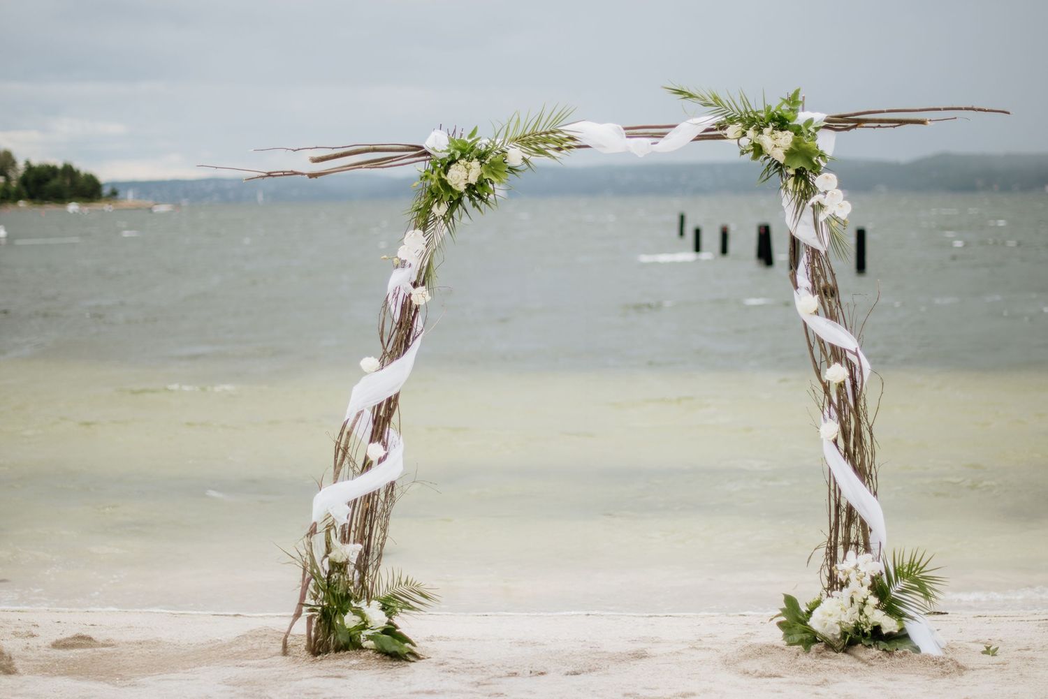 A rustic driftwood wedding arch decorated with white flowers stands on a beach overlooking the ocean at Villa Malla in Norway.