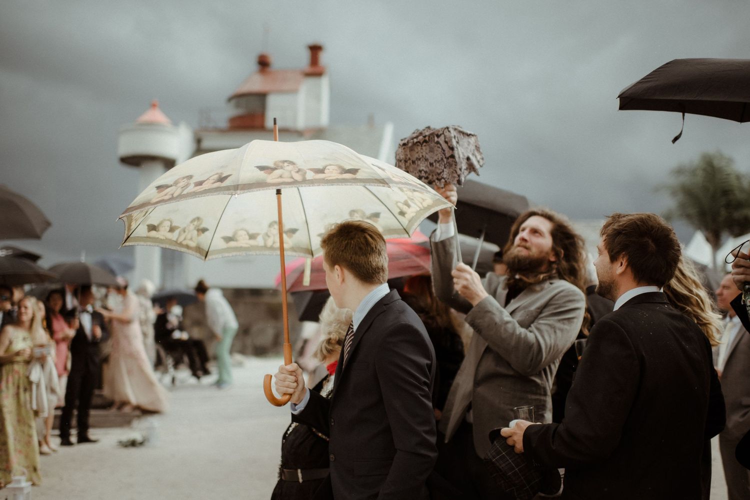 A group of wedding guests gather outdoors under a decorative umbrella on an overcast day during a celebration at Villa Malla in Norway.