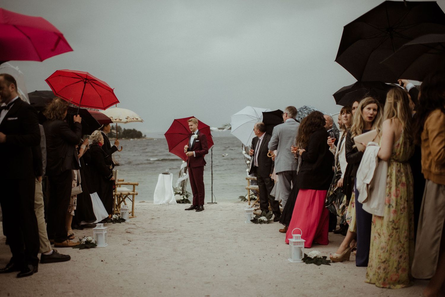 A beach wedding ceremony with guests holding red and black umbrellas on a cloudy day by the ocean at Villa Malla in Norway.