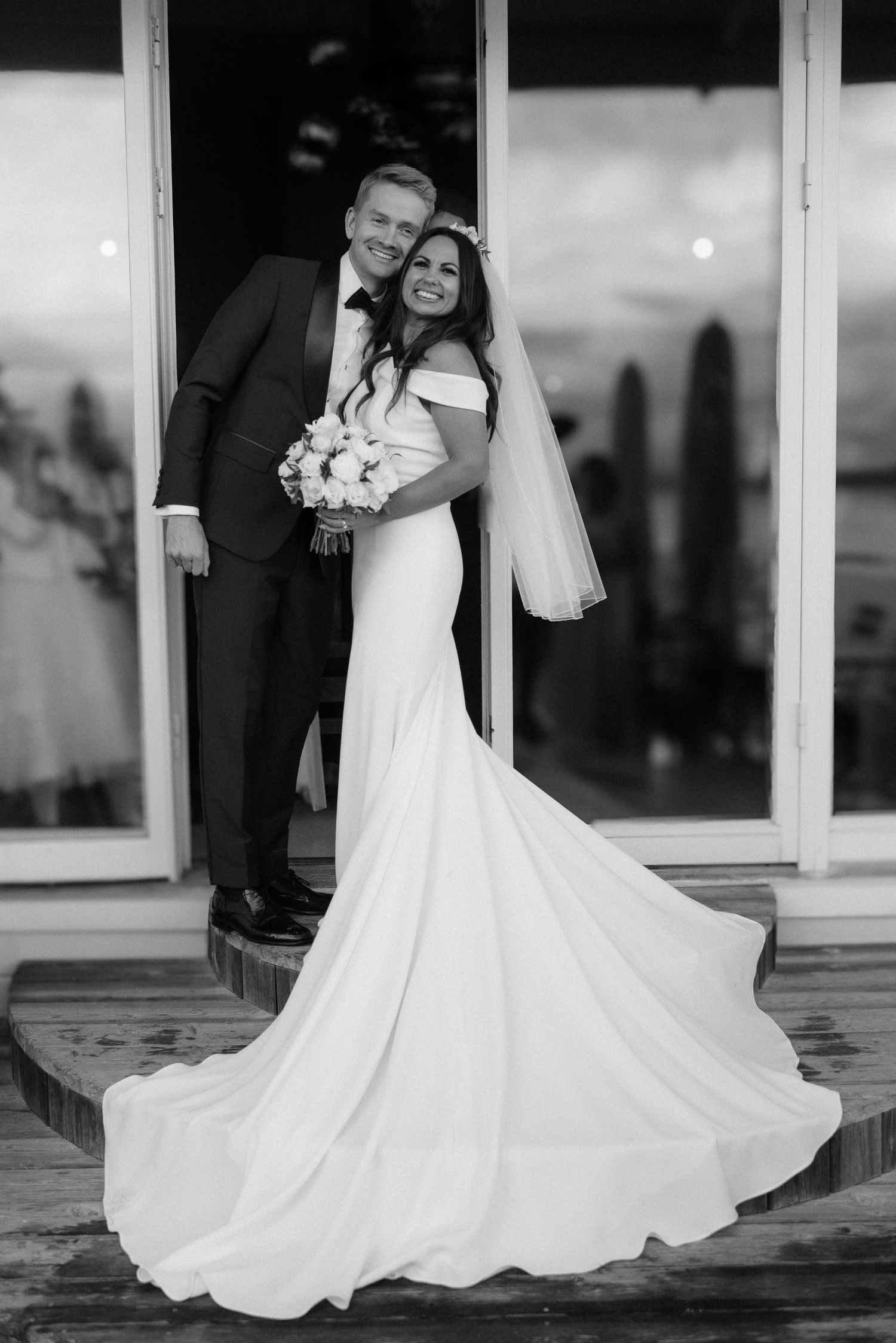 A black and white wedding photo featuring a couple in formal attire with a flowing white dress on a wooden deck at Villa Malla in Norway.