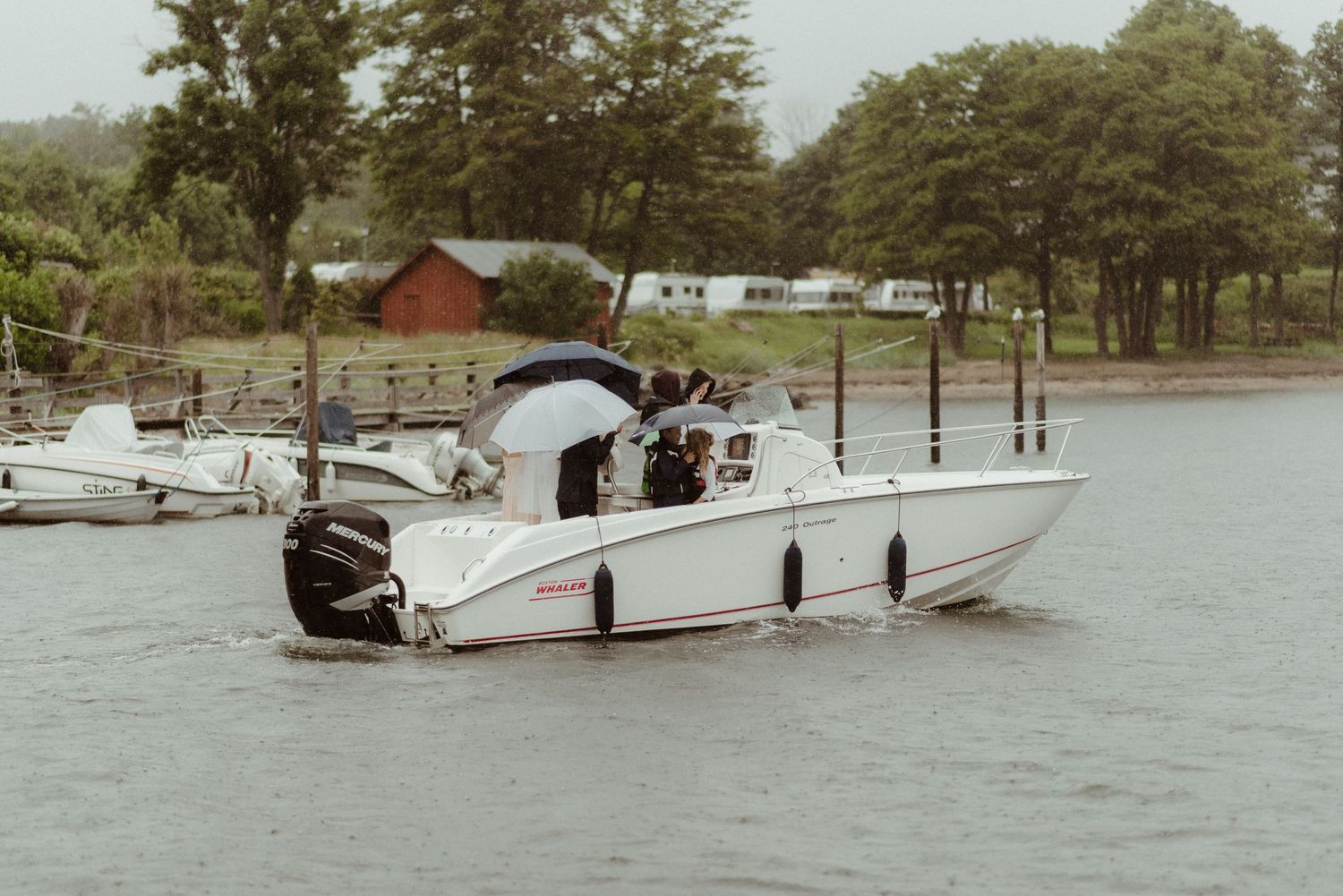 A white motorboat with passengers navigates along a lake shore with trees and a red barn in the background at Villa Malla in Norway.