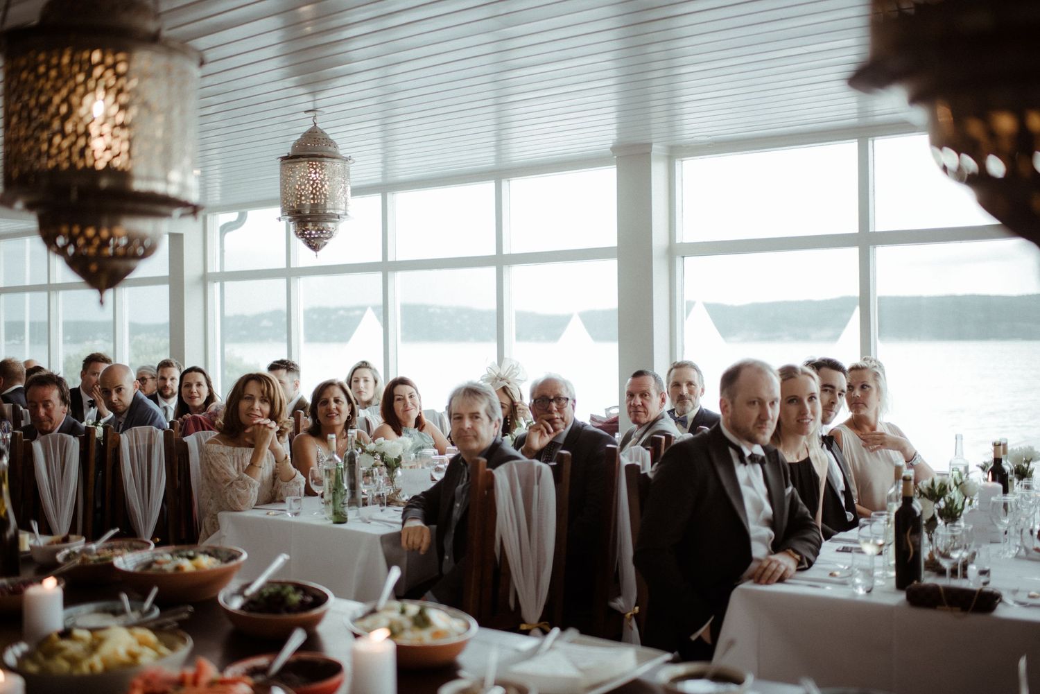 Wedding guests seated at tables celebrate during an elegant waterfront reception with hanging lantern decor at Villa Malla in Norway.